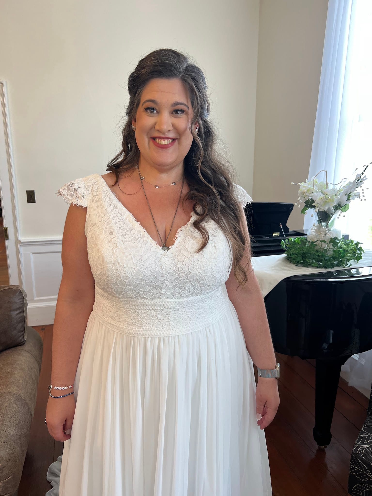 Smiling bride in a white lace cap-sleeve gown stands in an elegantly appointed room at Scarborough House with floral arrangements and a grand piano in the background.