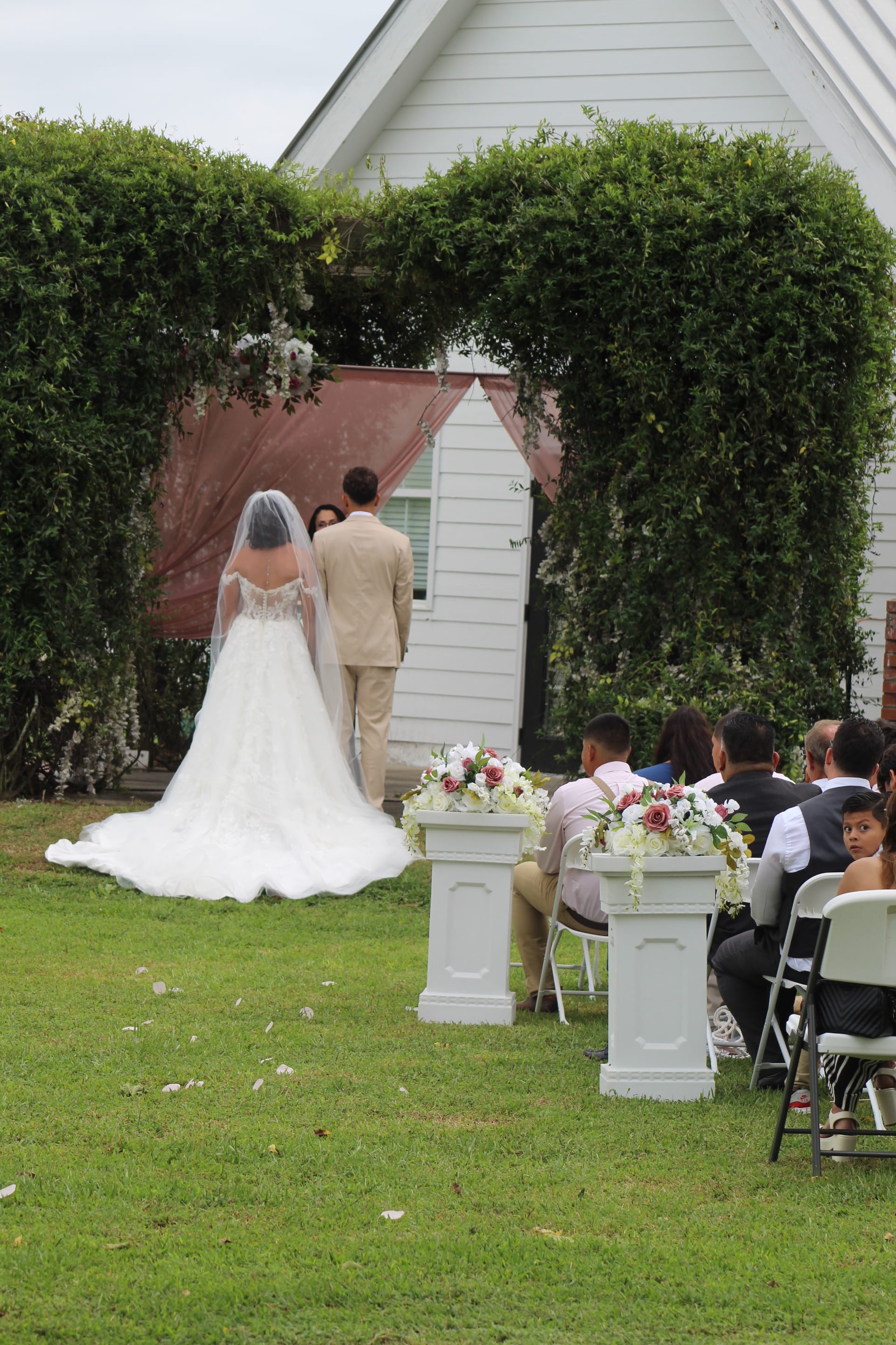 Bride in a white ball gown and veil stands with groom before an officiant under a lush green arch at an outdoor wedding ceremony on the lawn of a historic white estate chapel.