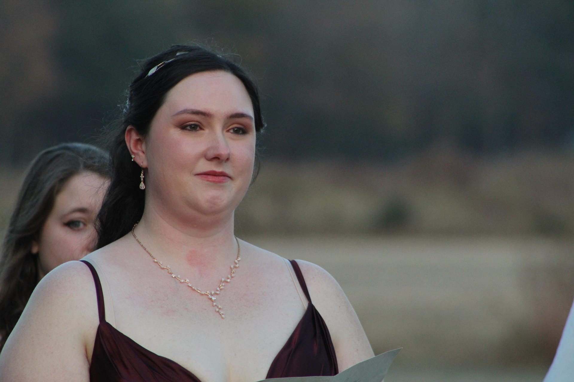 A bridesmaid in a burgundy gown and delicate pearl necklace stands on the ceremony lawn at Scarborough House, holding papers with a thoughtful expression during the wedding ceremony.