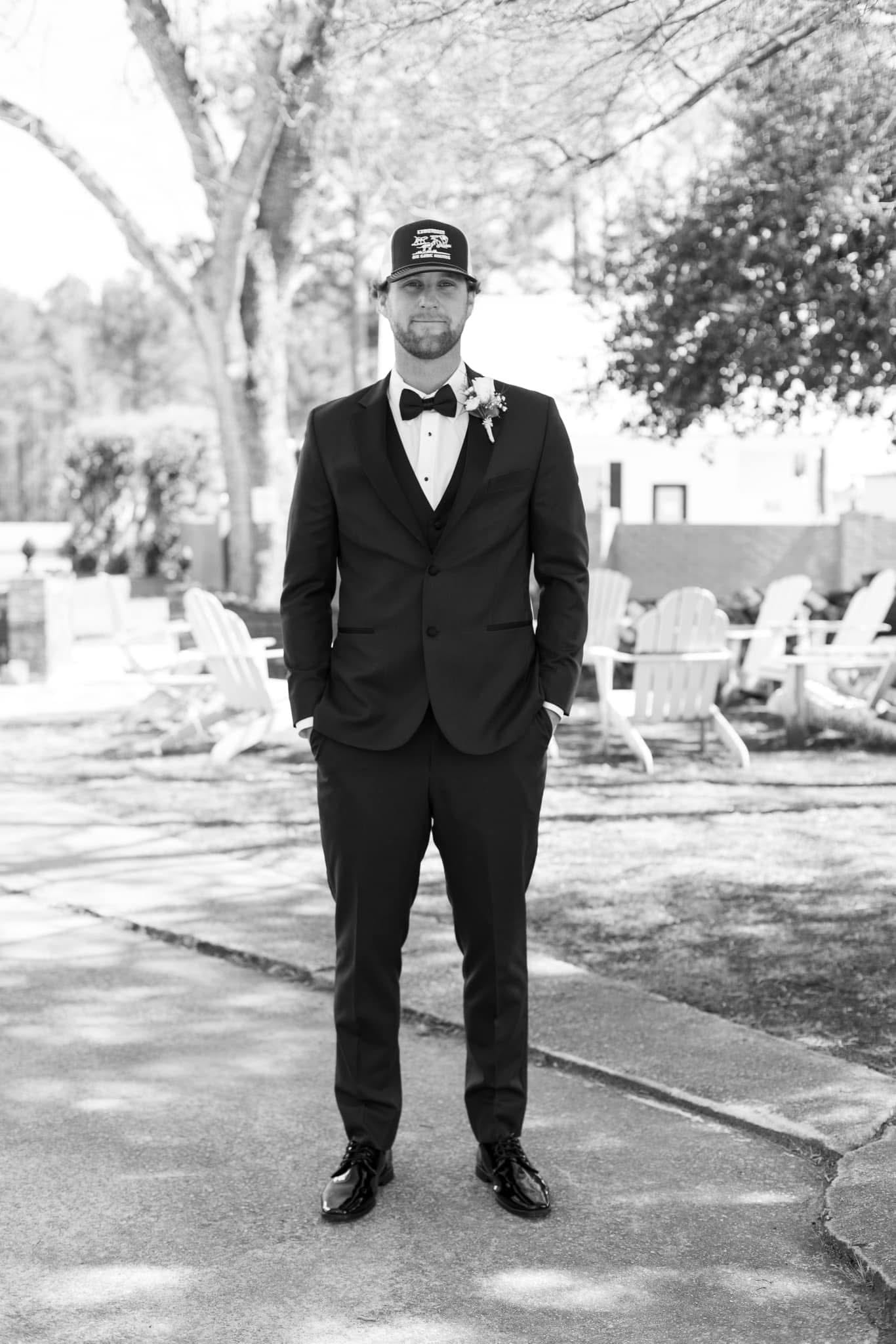 Groom in a black tuxedo with bow tie and boutonniere wearing a baseball cap stands confidently on the stone pathway at Scarborough House with Adirondack chairs and oak trees in the background.