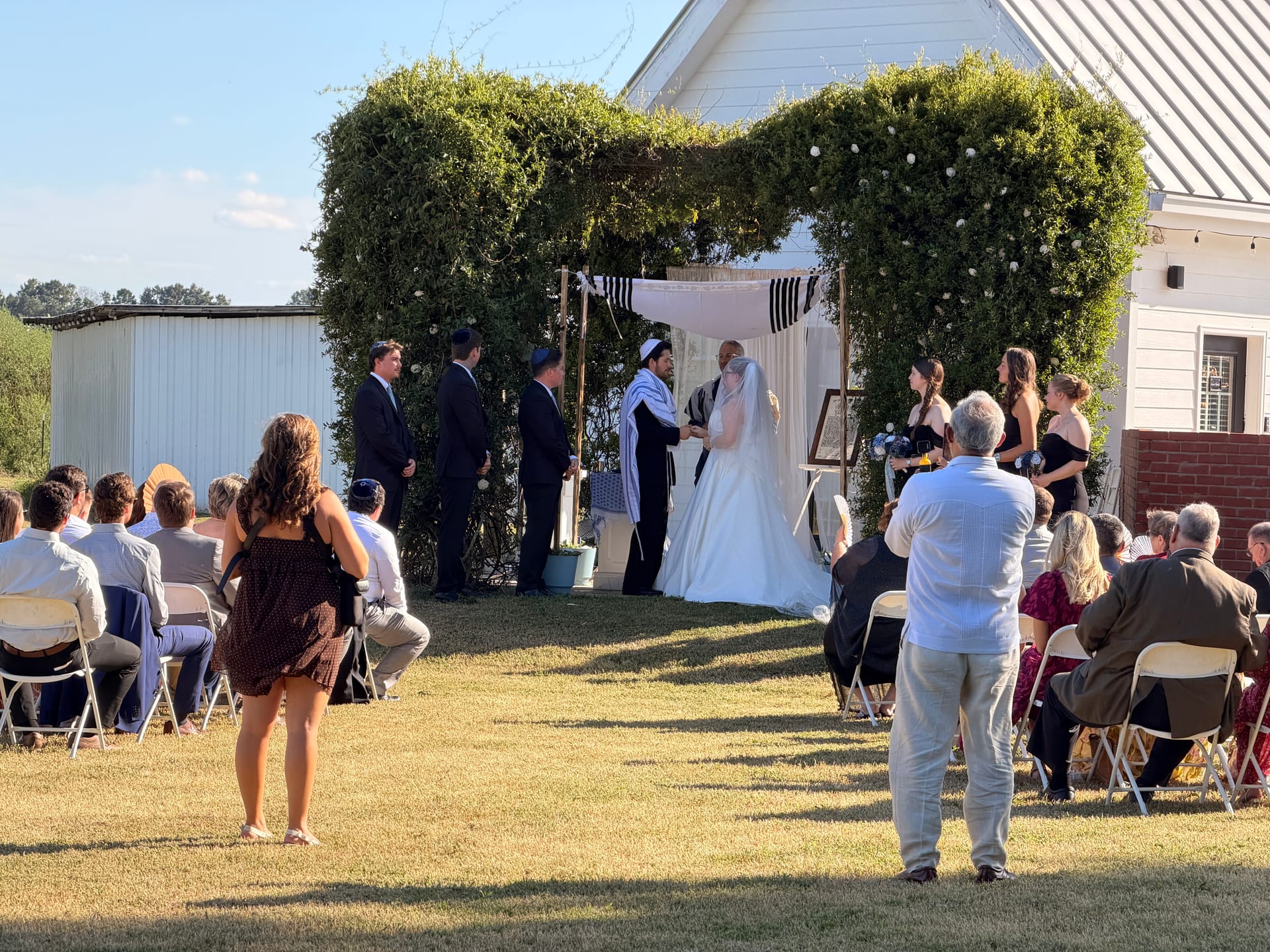 Bride and groom exchange vows beneath a lush green chuppah draped in white fabric on the sunlit ceremony lawn at Scarborough House historic estate wedding venue.