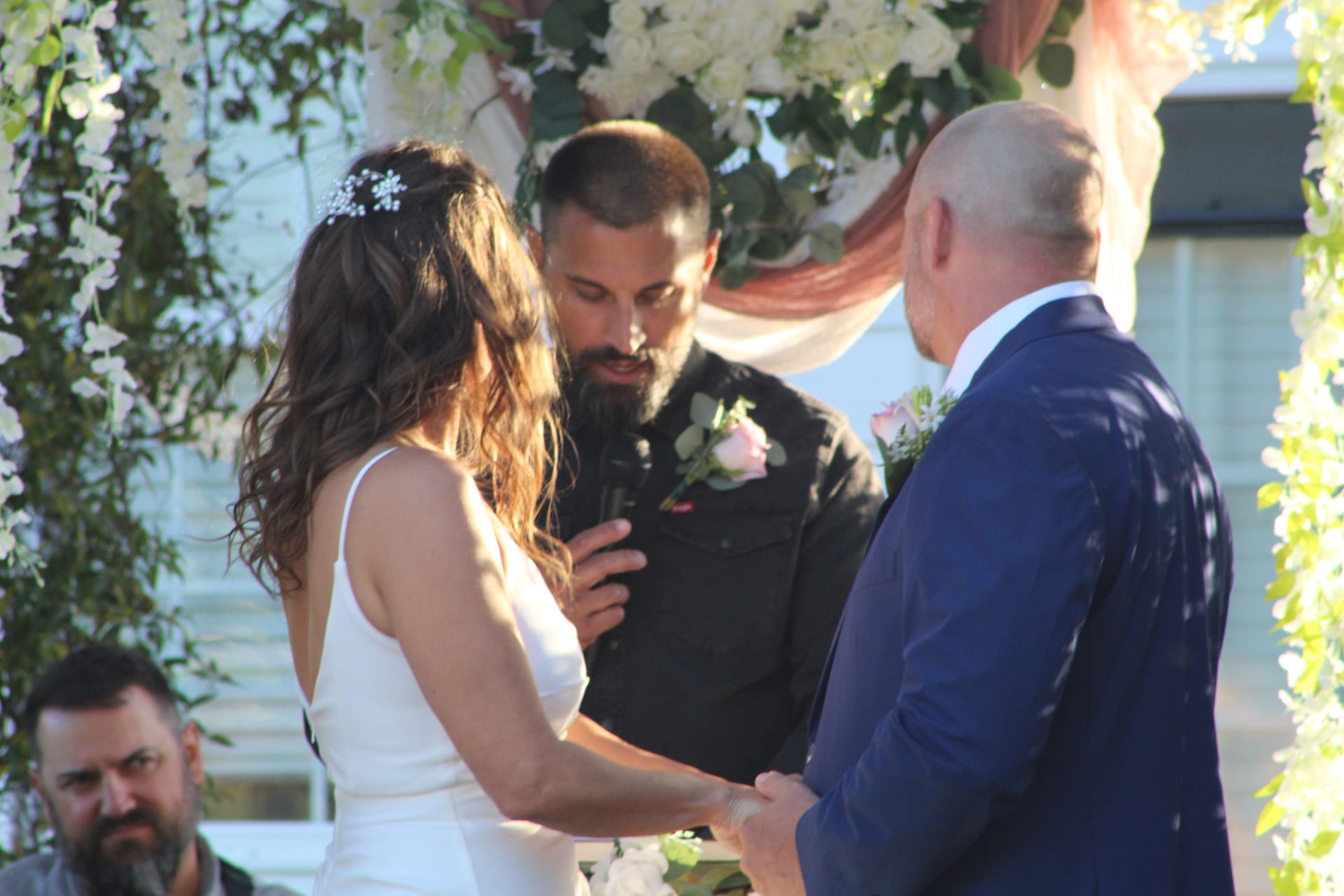 Bride and groom exchange vows beneath a lush white floral and greenery arch with blush draping at Scarborough House outdoor ceremony.