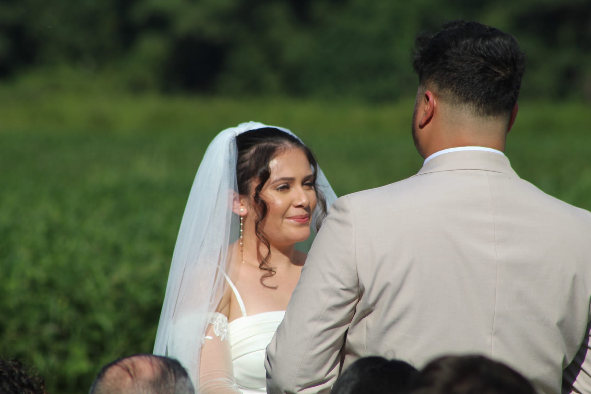 Bride in a white gown and veil gazes emotionally at her groom during an outdoor wedding ceremony at Scarborough House, with lush green fields and trees in the background.