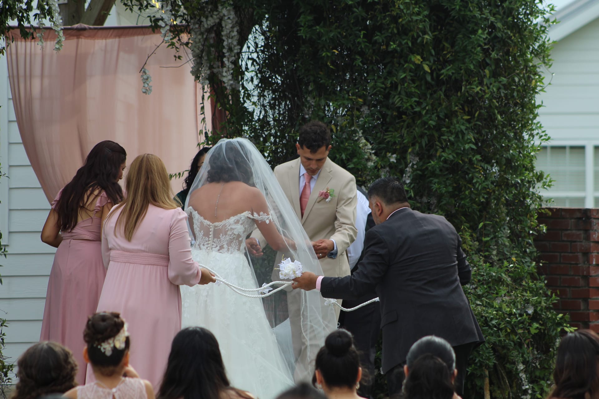 Bride and groom participate in a lasso unity cord ceremony ritual surrounded by bridesmaids in blush pink gowns beneath a draped floral altar at an outdoor garden wedding.