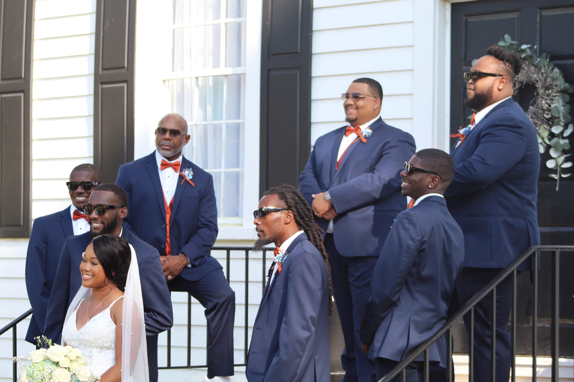 Bride in white gown holding bouquet stands with groomsmen in navy suits and orange bow ties on the front steps of the historic 1821 Scarborough House manor.