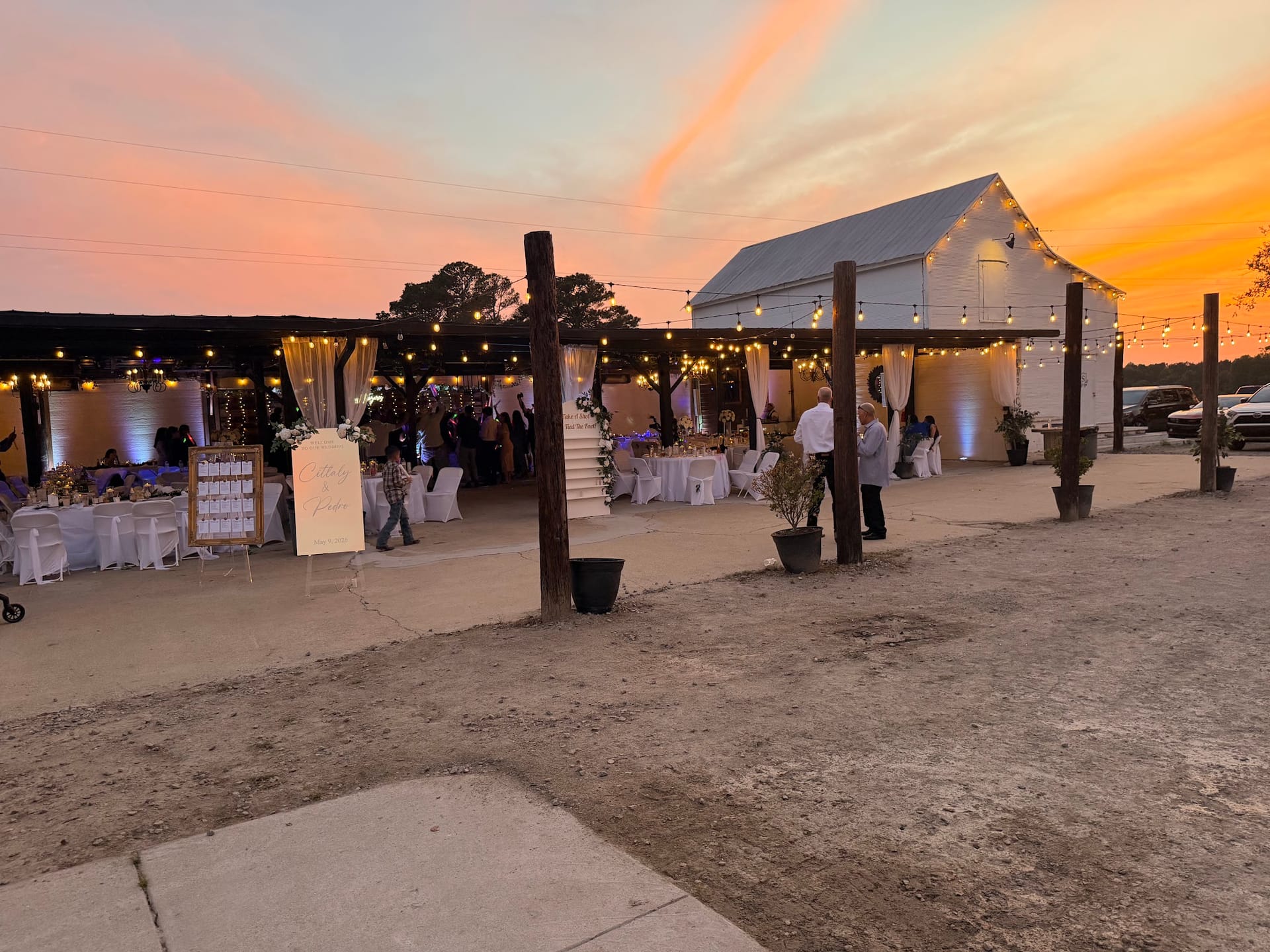 A warmly lit outdoor wedding reception at Scarborough House glows beneath a dramatic pink and orange sunset sky with string lights draped between rustic wooden posts and white-draped tables filled with guests.