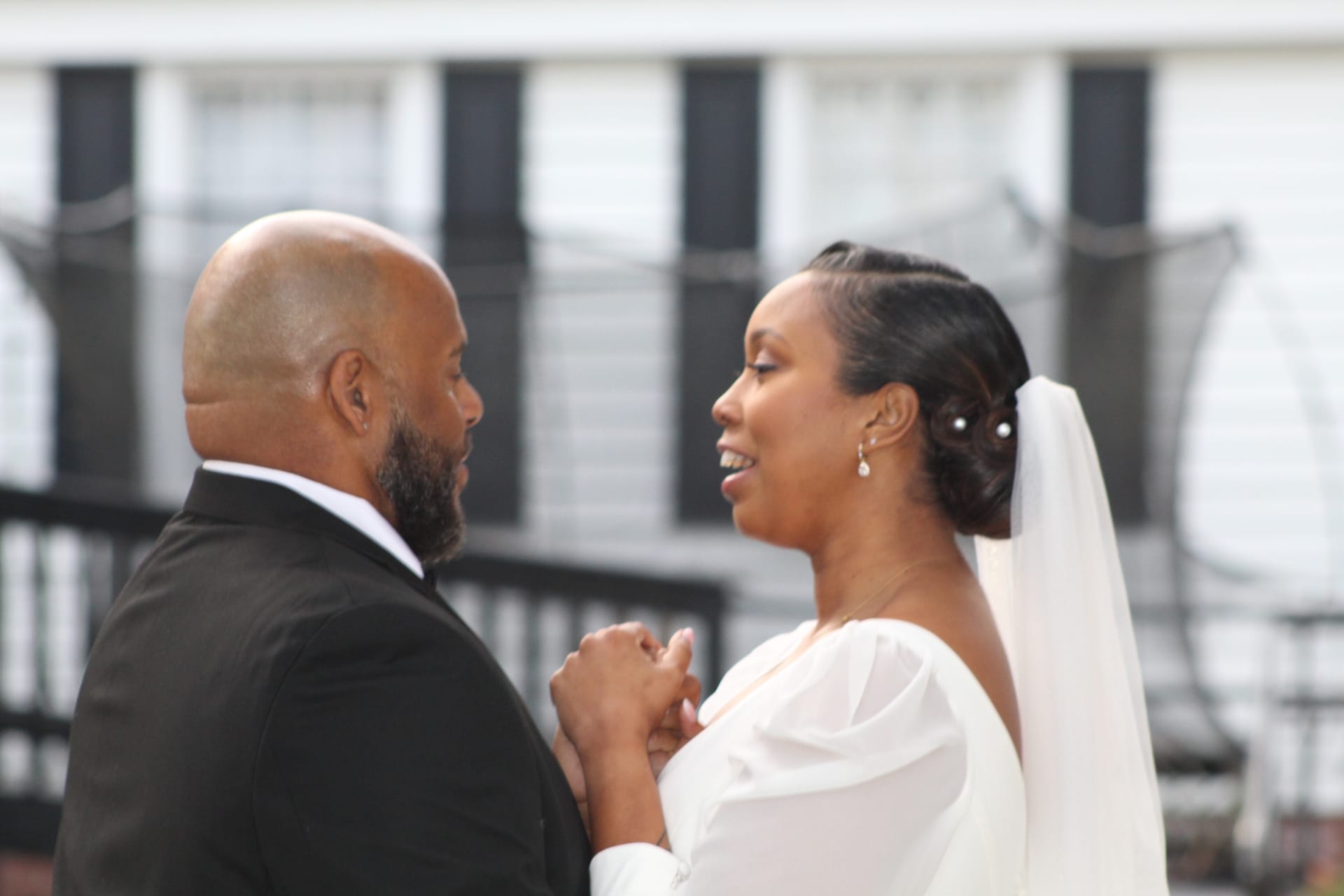 Bride in a white off-shoulder gown and veil smiles warmly at her groom in a black tuxedo as they hold hands in front of the historic Scarborough House manor.