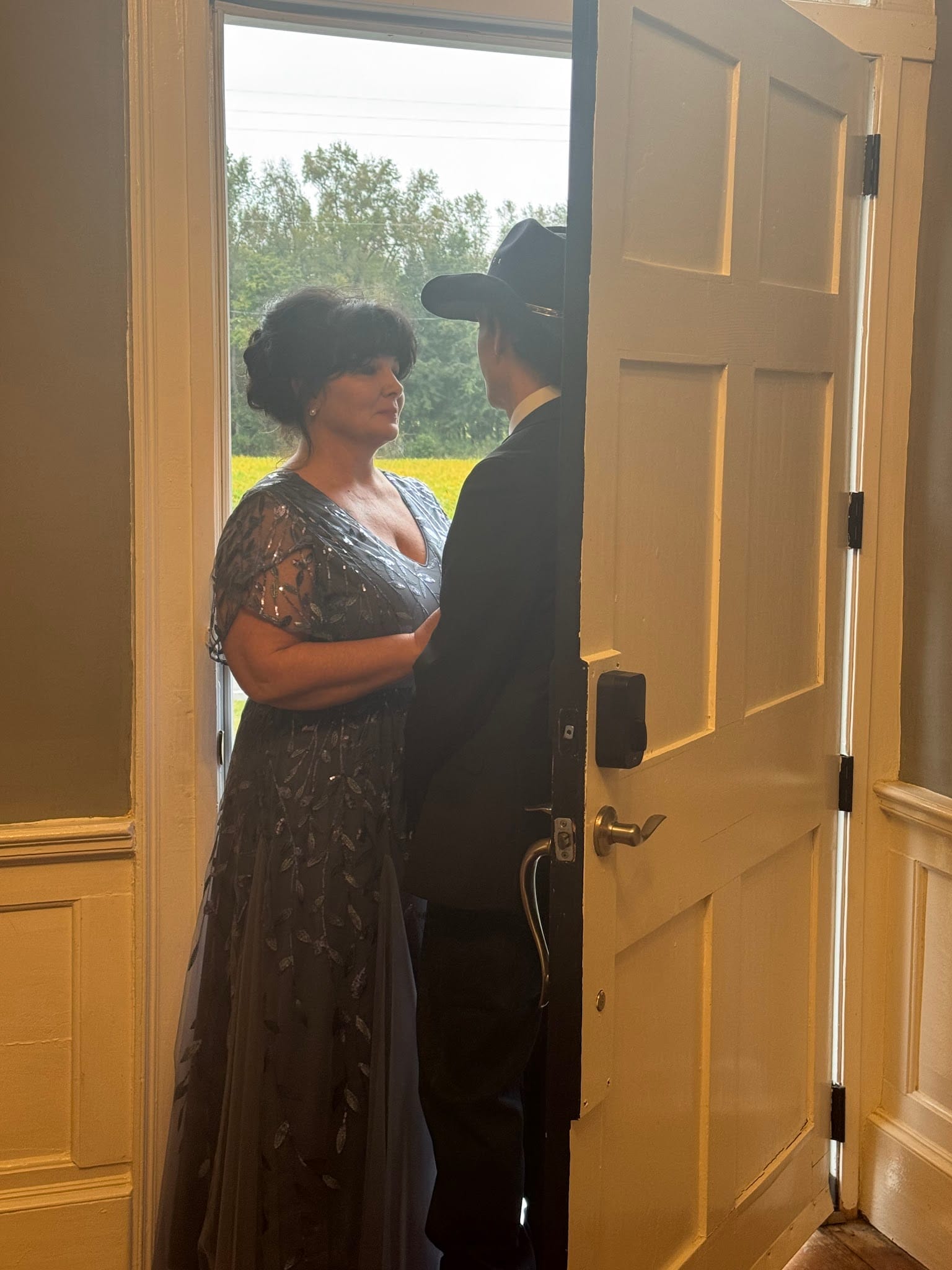 A woman in a sparkling blue-grey gown faces a man in a black cowboy hat through the open white door of the 1821 Scarborough House manor, with golden sunlight and lush green fields visible behind them.