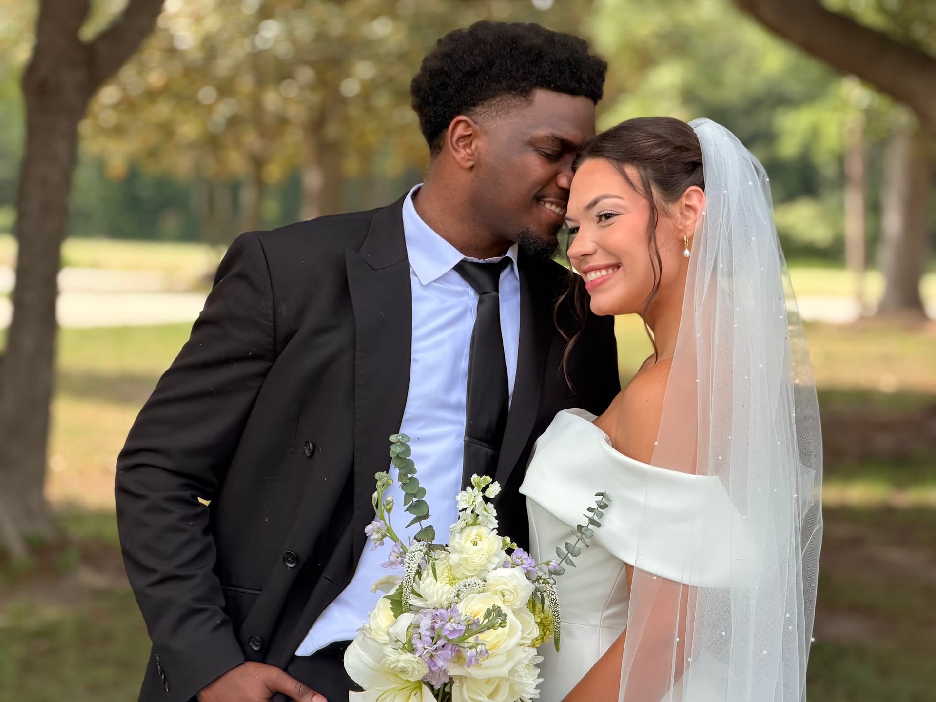 Bride in off-shoulder white gown and veil smiles as groom in black suit tenderly leans in close, holding a bouquet of white and lavender flowers beneath the oak trees at Scarborough House.