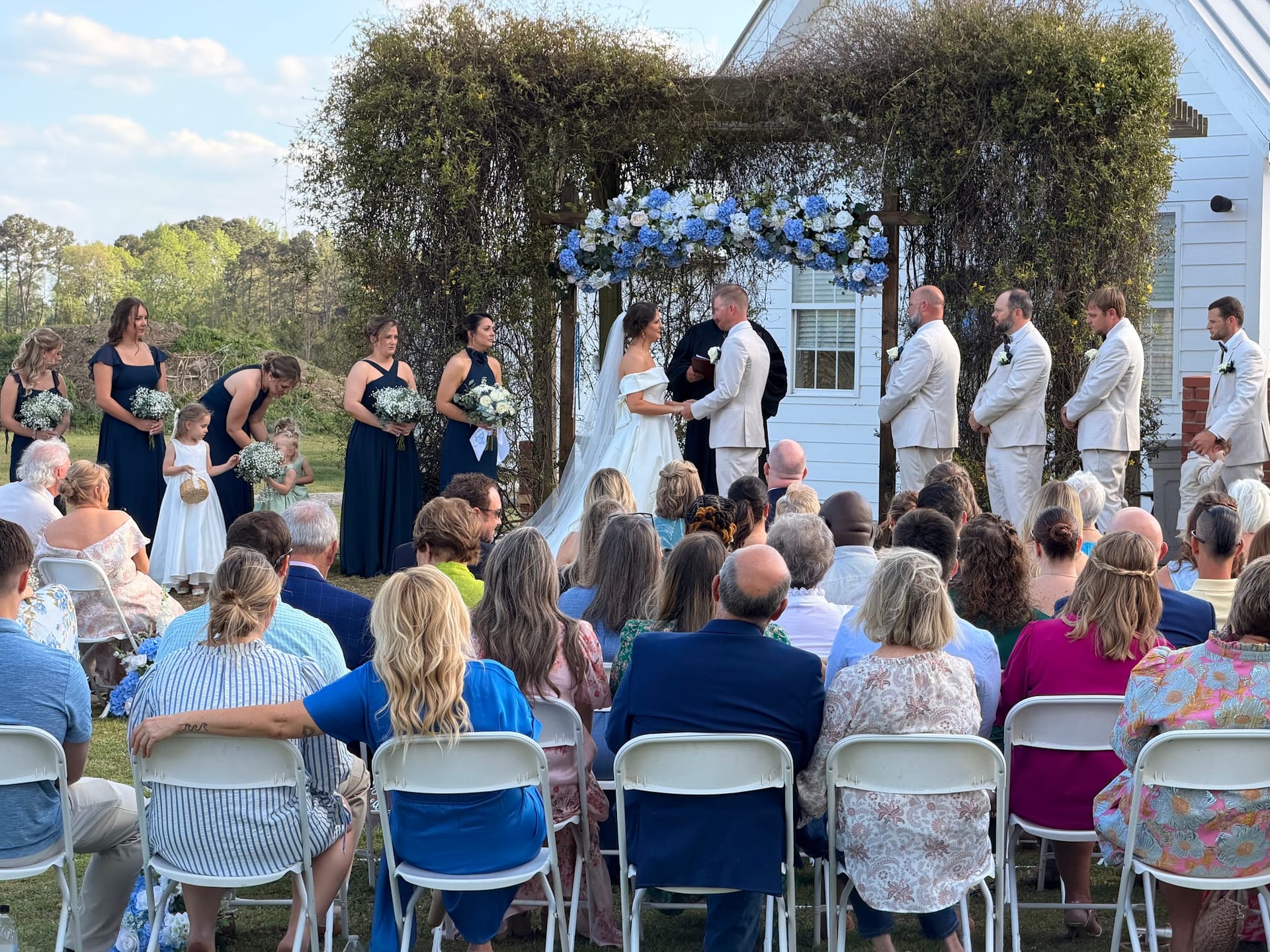 Outdoor wedding ceremony at Scarborough House with bride and groom exchanging vows beneath a lush greenery arch adorned with blue and white florals, surrounded by bridesmaids in navy, groomsmen in tan, and seated guests on a sunlit lawn.