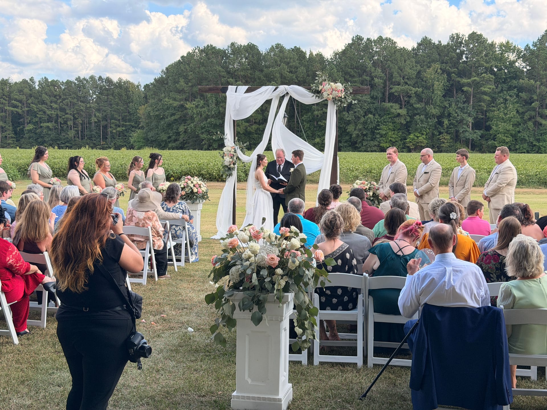 Outdoor wedding ceremony on the Scarborough House ceremony lawn with a draped wooden arch adorned with floral arrangements, a bride and groom exchanging vows before guests seated in white chairs surrounded by farmland fields and lush trees.