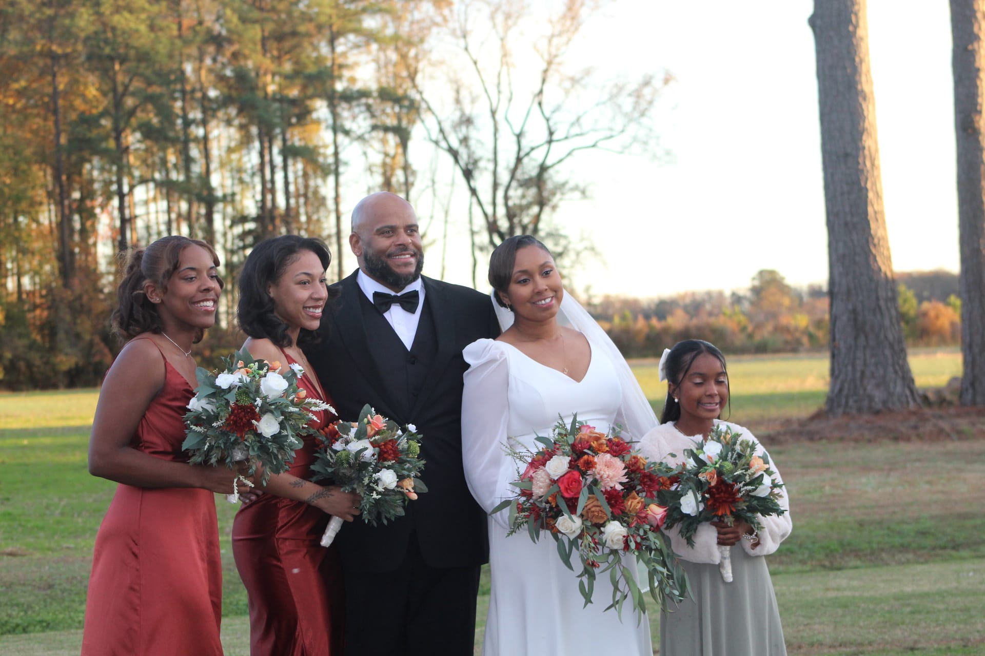 Bride in a white gown and veil poses with groom in a black tuxedo and two bridesmaids in rust-colored dresses and a young girl in sage, all holding lush autumn floral bouquets on the estate lawn surrounded by golden fall foliage.