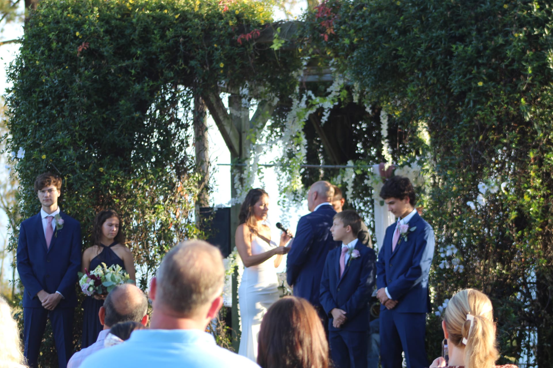 Bride and groom exchanging vows beneath a lush ivy-draped archway on the ceremony lawn at Scarborough House, surrounded by family and white floral accents.