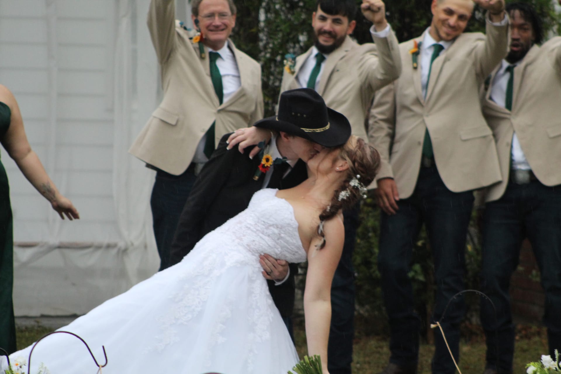 Bride in white lace gown and groom in black jacket and cowboy hat share a dramatic dip kiss while groomsmen in tan blazers cheer behind them at an outdoor wedding ceremony.