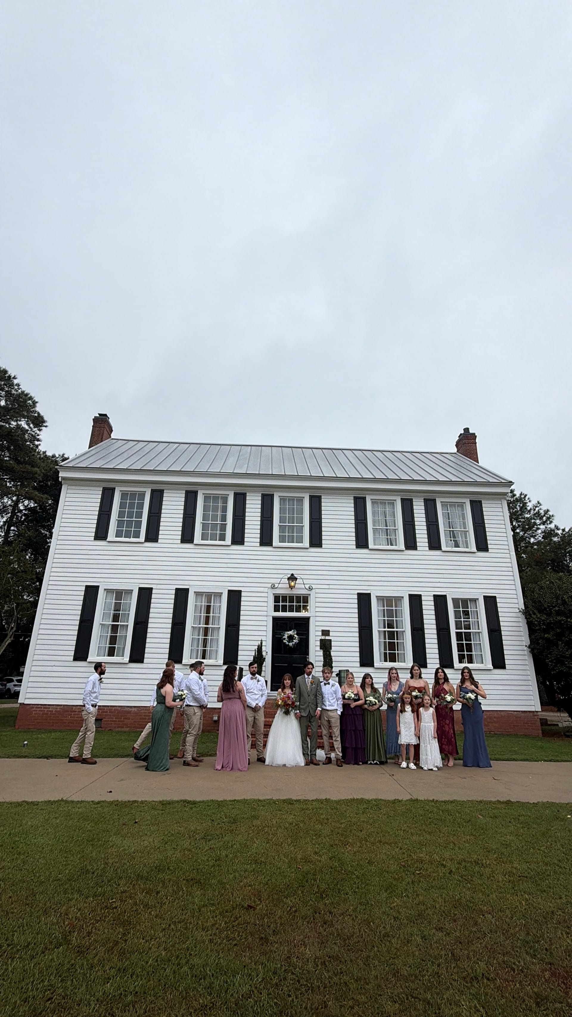 Wedding party gathered in front of the white historic Scarborough House manor on an overcast day, bridesmaids in jewel-tone gowns flanking the bride and groom at the main entrance.