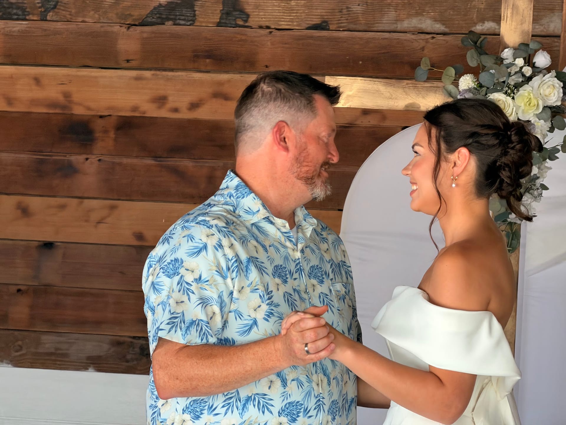 Bride in an off-shoulder white gown and groom in a blue floral shirt share a tender first dance in front of a rustic wood wall and white floral arch at Scarborough House.