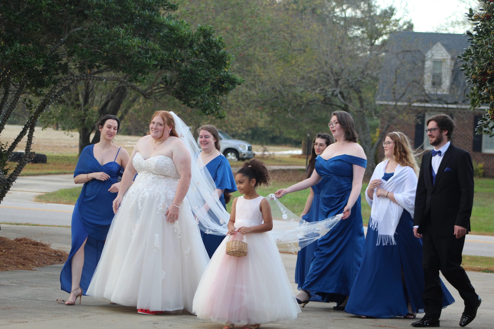 Smiling bride in a strapless lace ballgown and veil walks with her bridal party in royal-blue gowns and a flower girl in blush on the grounds of a historic estate wedding venue.