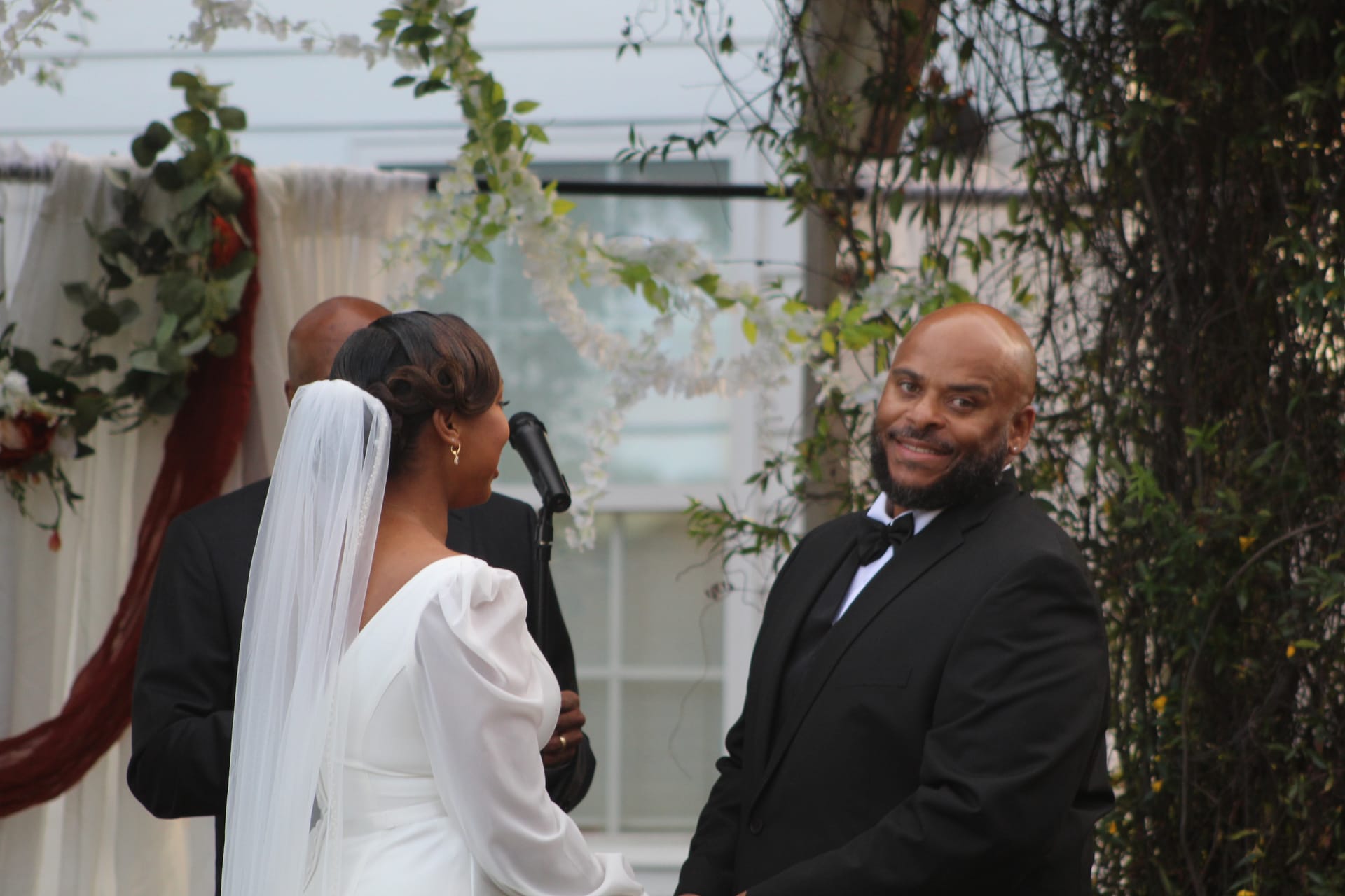 Groom in black tuxedo smiling warmly at his bride in a white gown and veil during an outdoor wedding ceremony beneath a floral arch at Scarborough House.
