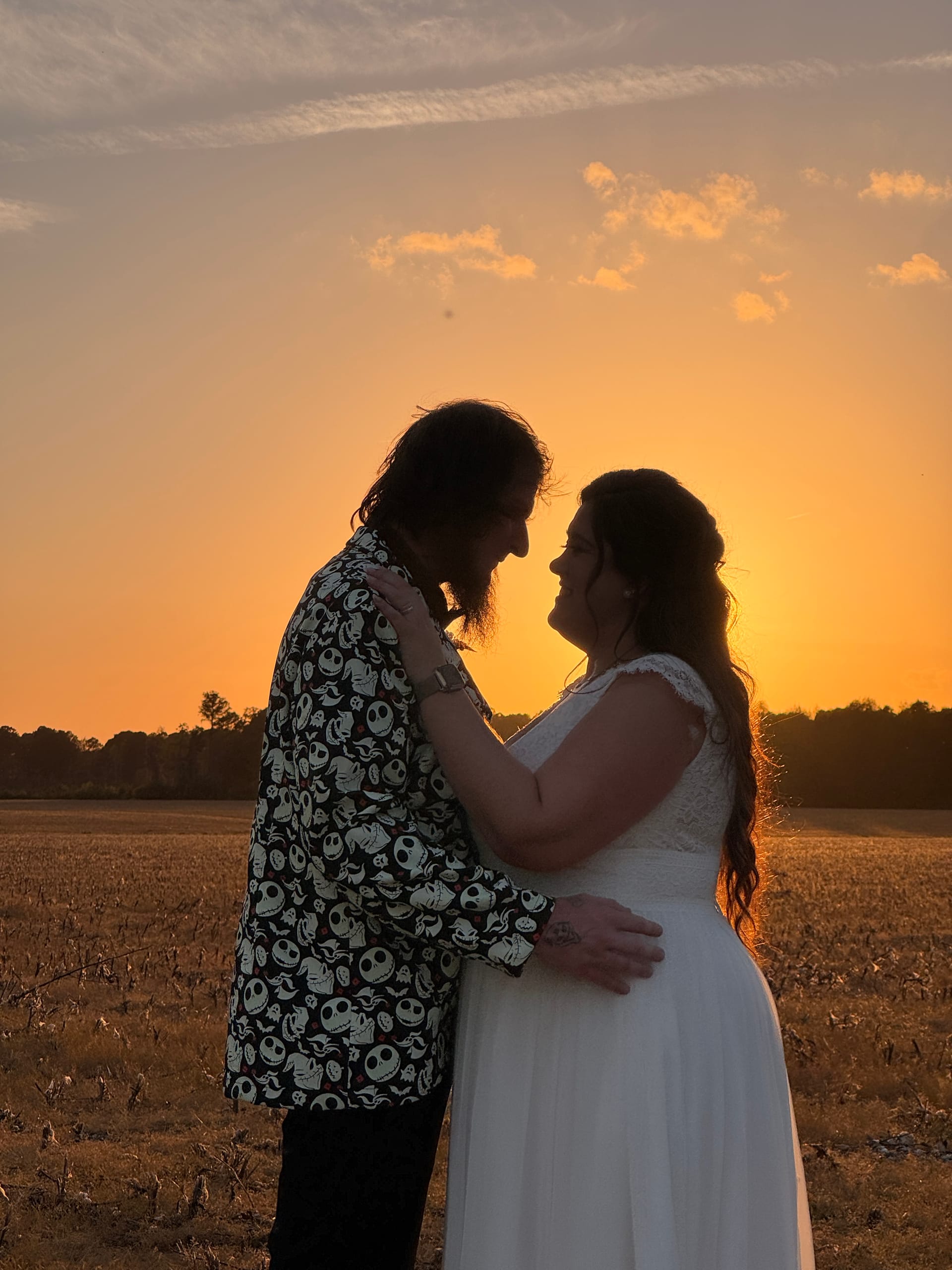 Bride in white lace gown and groom in a Nightmare Before Christmas printed shirt share a romantic near-kiss silhouette against a glowing orange sunset over open farmland fields at Scarborough House.