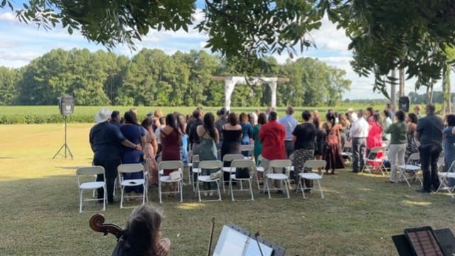 Guests standing and gathered at an outdoor wedding ceremony on the lawn at Scarborough House with a white arbor surrounded by green fields and mature trees in the background