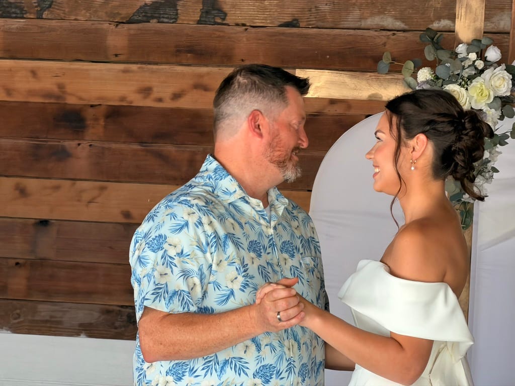 A smiling bride in an off-shoulder white gown holds hands with her groom in a floral shirt beneath a circular floral arch decorated with white roses and eucalyptus at Scarborough House wedding venue.