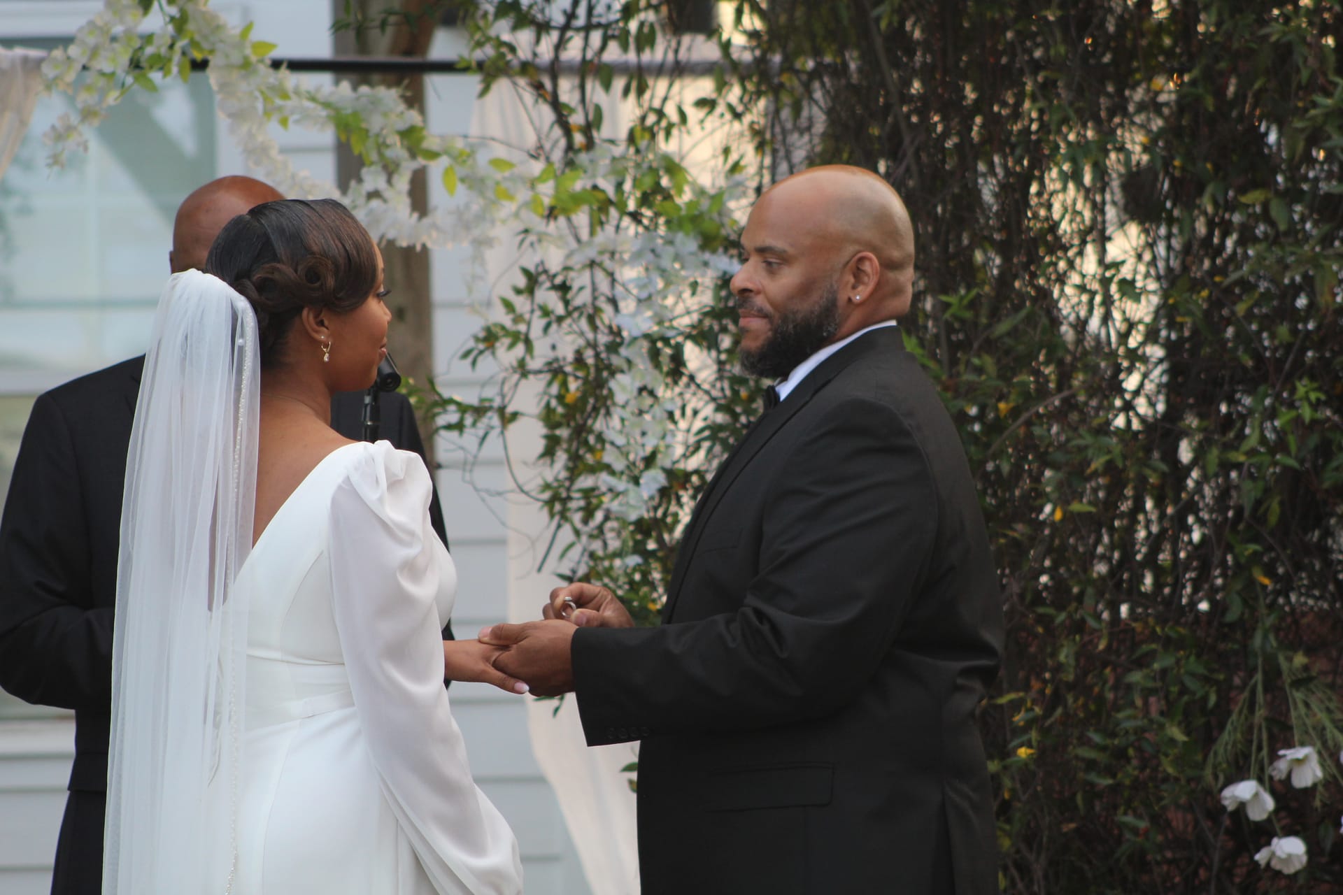 Groom smiling warmly while placing a ring on the bride's hand during an outdoor wedding ceremony at a historic estate surrounded by lush greenery.
