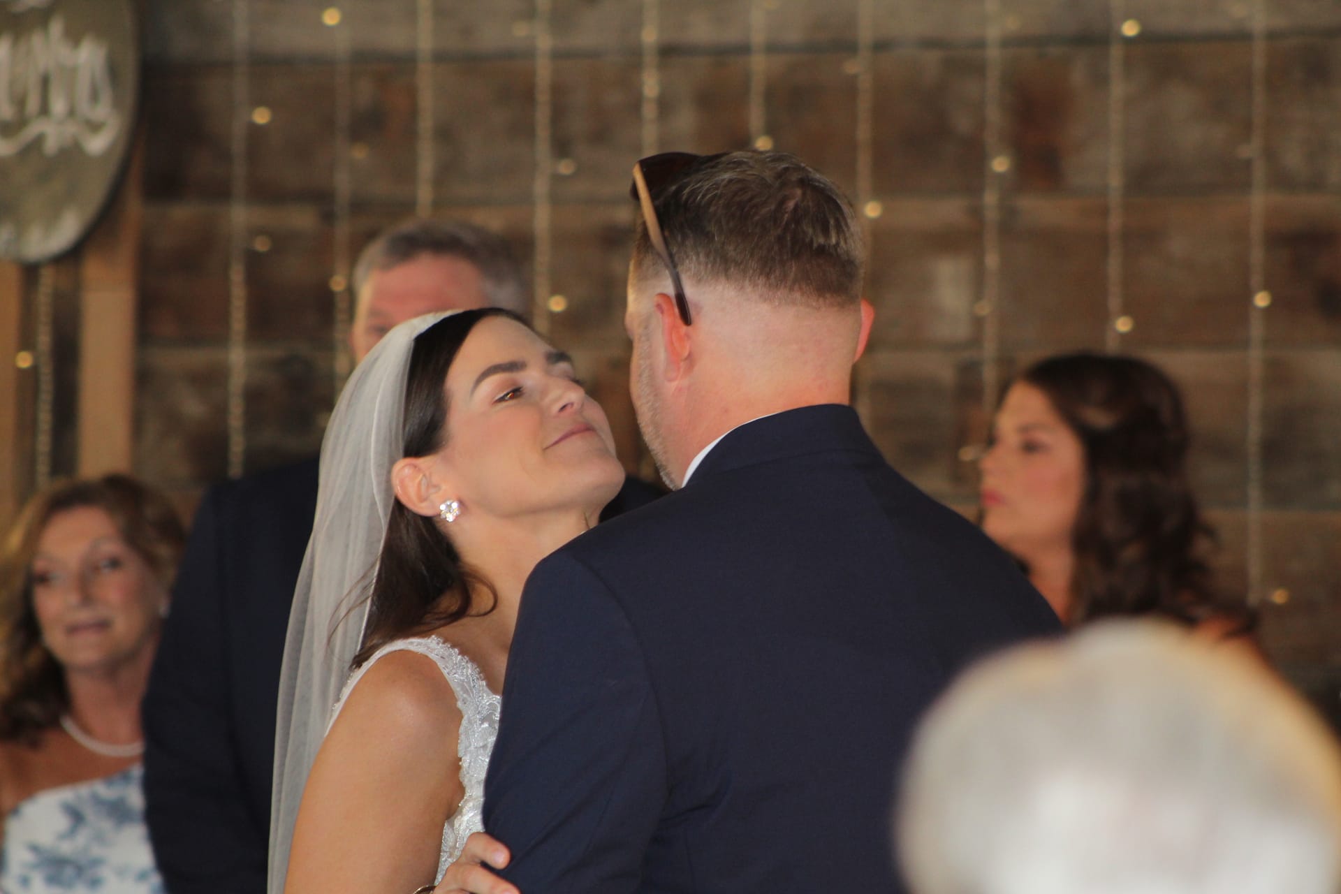 Bride in a lace gown and veil gazes tenderly up at her groom during their wedding ceremony inside a warmly lit barn-style venue.