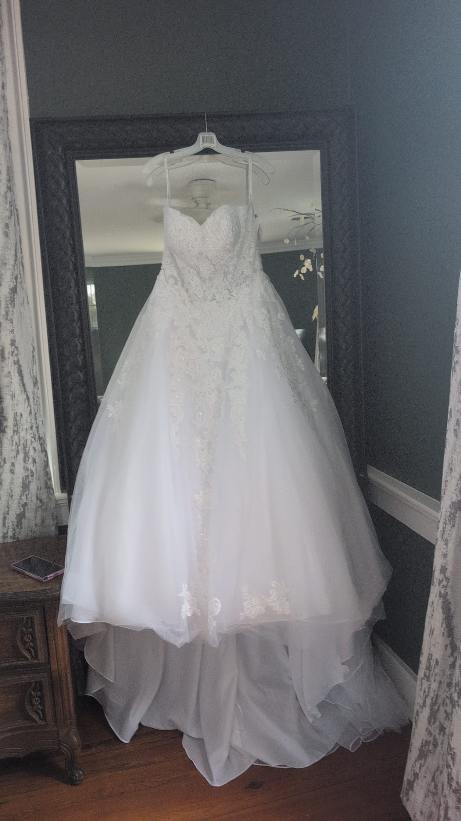 A strapless white ball-gown wedding dress with lace appliqué bodice and layered tulle skirt hanging on a white hanger in front of a large ornate dark-framed mirror inside Scarborough House.