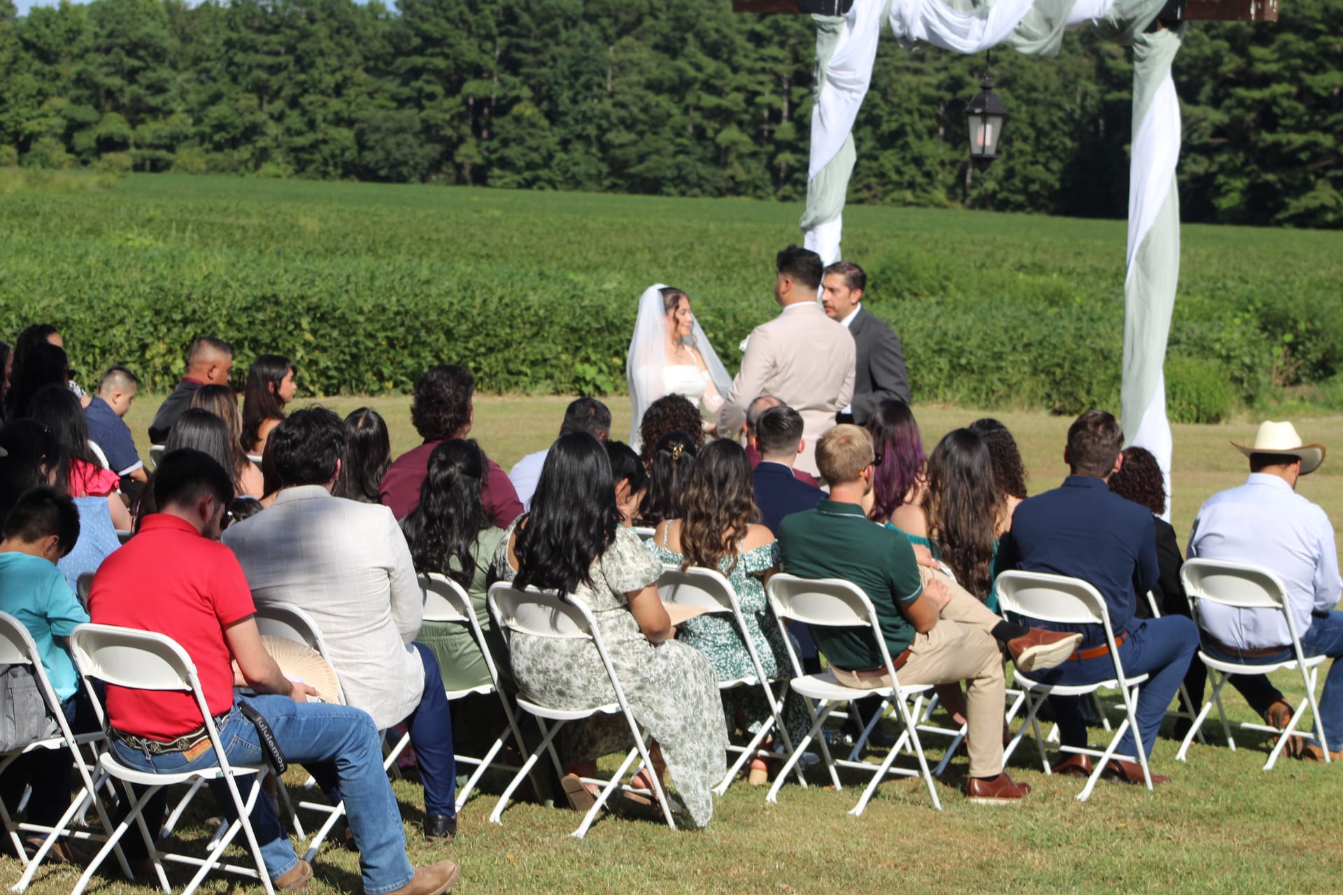 Guests seated in white folding chairs on the ceremony lawn watch a bride in a white veil and groom exchange vows beneath a draped arch, surrounded by green farmland fields and wooded treeline at Scarborough House.
