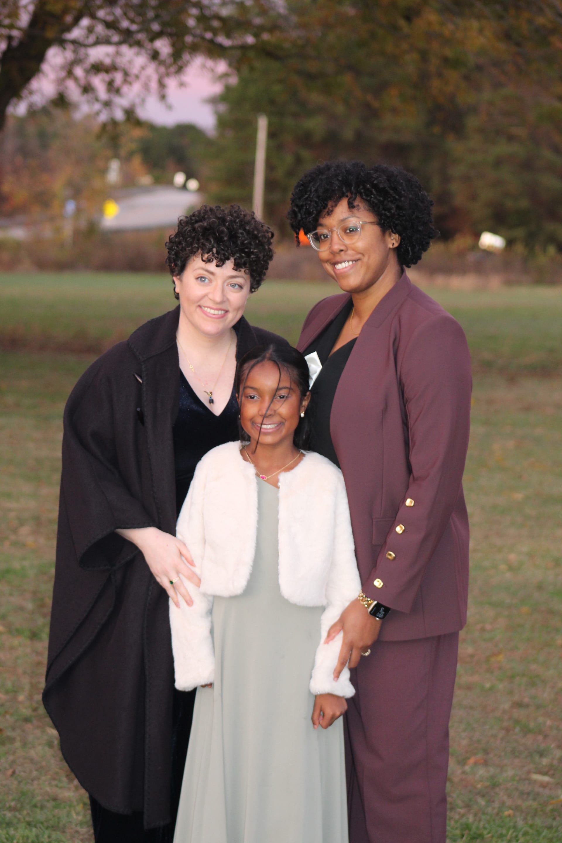 Two women and a young girl dressed in formal attire smile warmly together on the ceremony lawn at Scarborough House during an autumn wedding celebration.