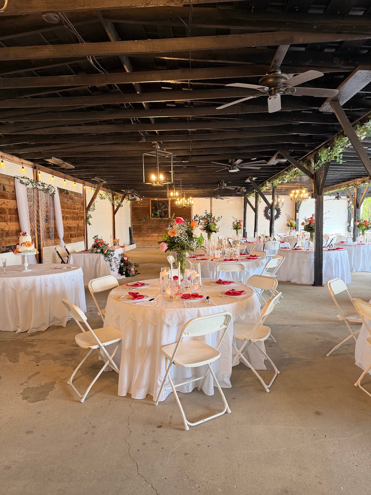 Elegantly decorated reception tables with white linens, red napkins, and colorful floral centerpieces beneath a rustic wood-beamed pavilion with chandeliers and ceiling fans at Scarborough House.