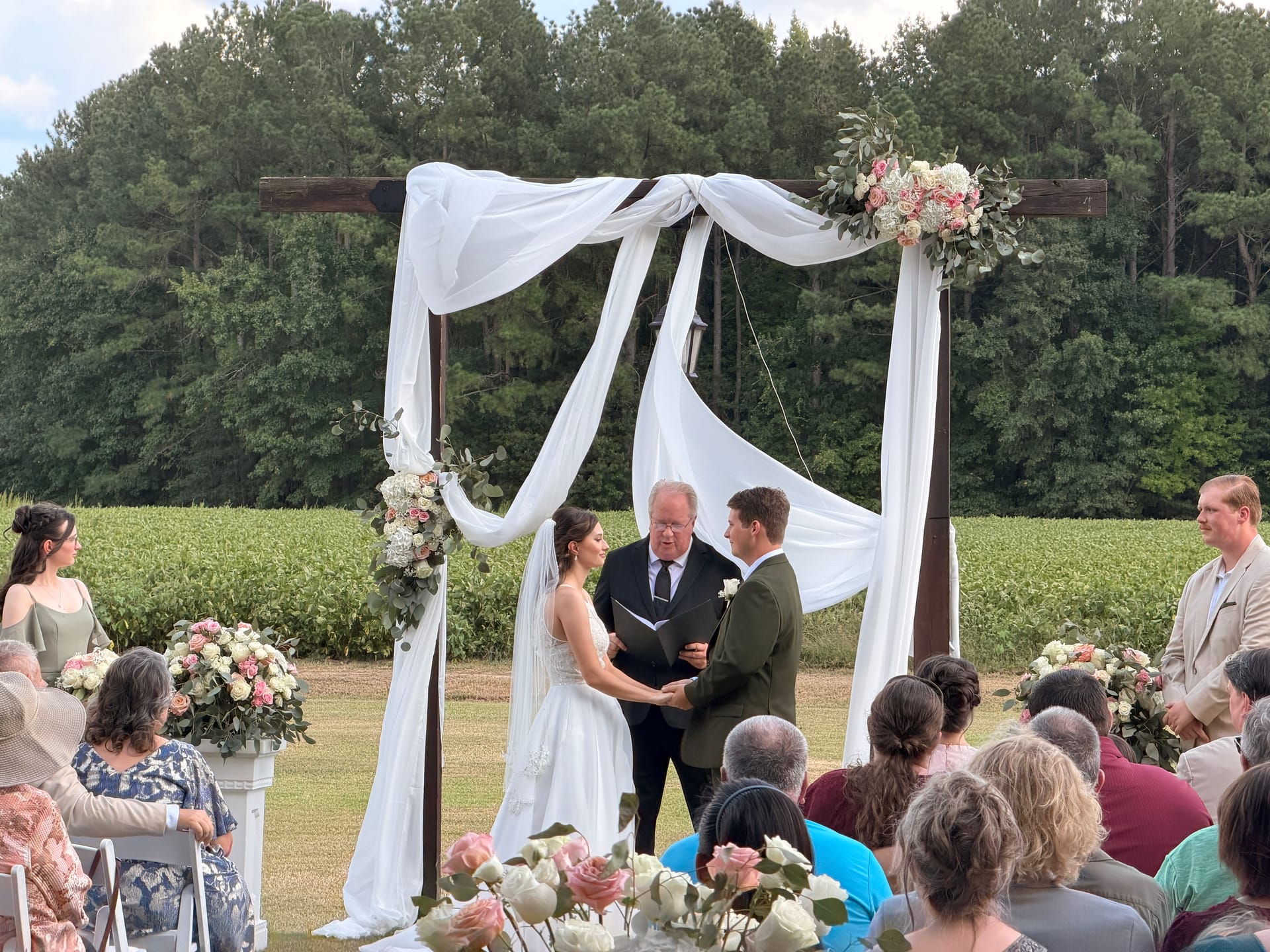 Bride and groom exchange vows beneath a draped wooden arch adorned with pink and white florals on the ceremony lawn at Scarborough House, surrounded by guests with rolling farmland fields in the background.