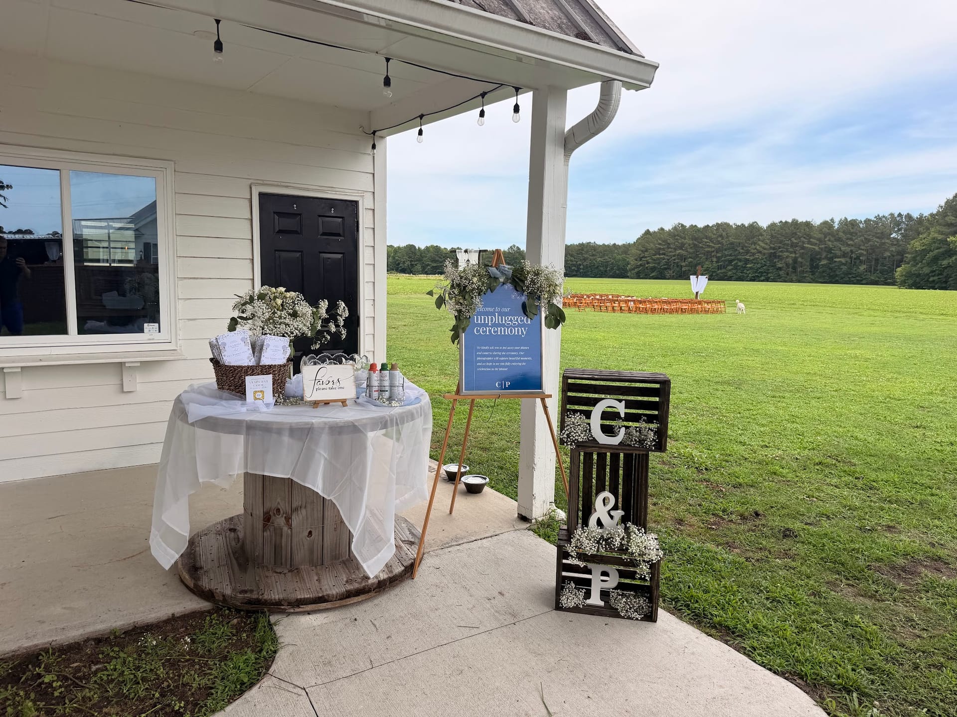 Wedding welcome table draped in white linen atop a wooden spool reel on the venue porch, flanked by an unplugged ceremony sign on an easel and stacked wooden crates monogrammed with C and P, with the ceremony lawn visible in the background.