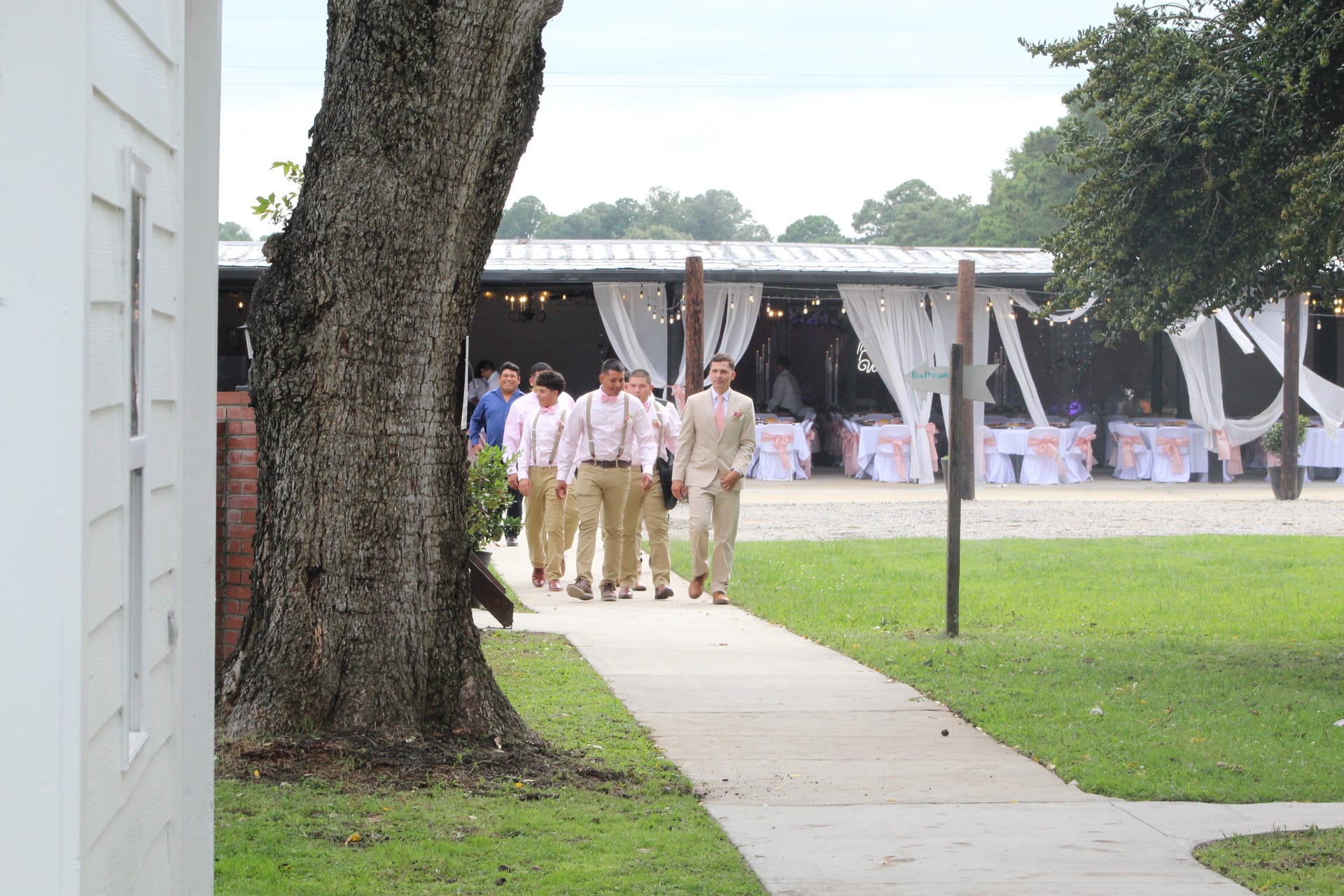 Groom in a tan suit leads his groomsmen in matching pink shirts and khaki pants along a shaded walkway toward the decorated outdoor reception pavilion at Scarborough House.
