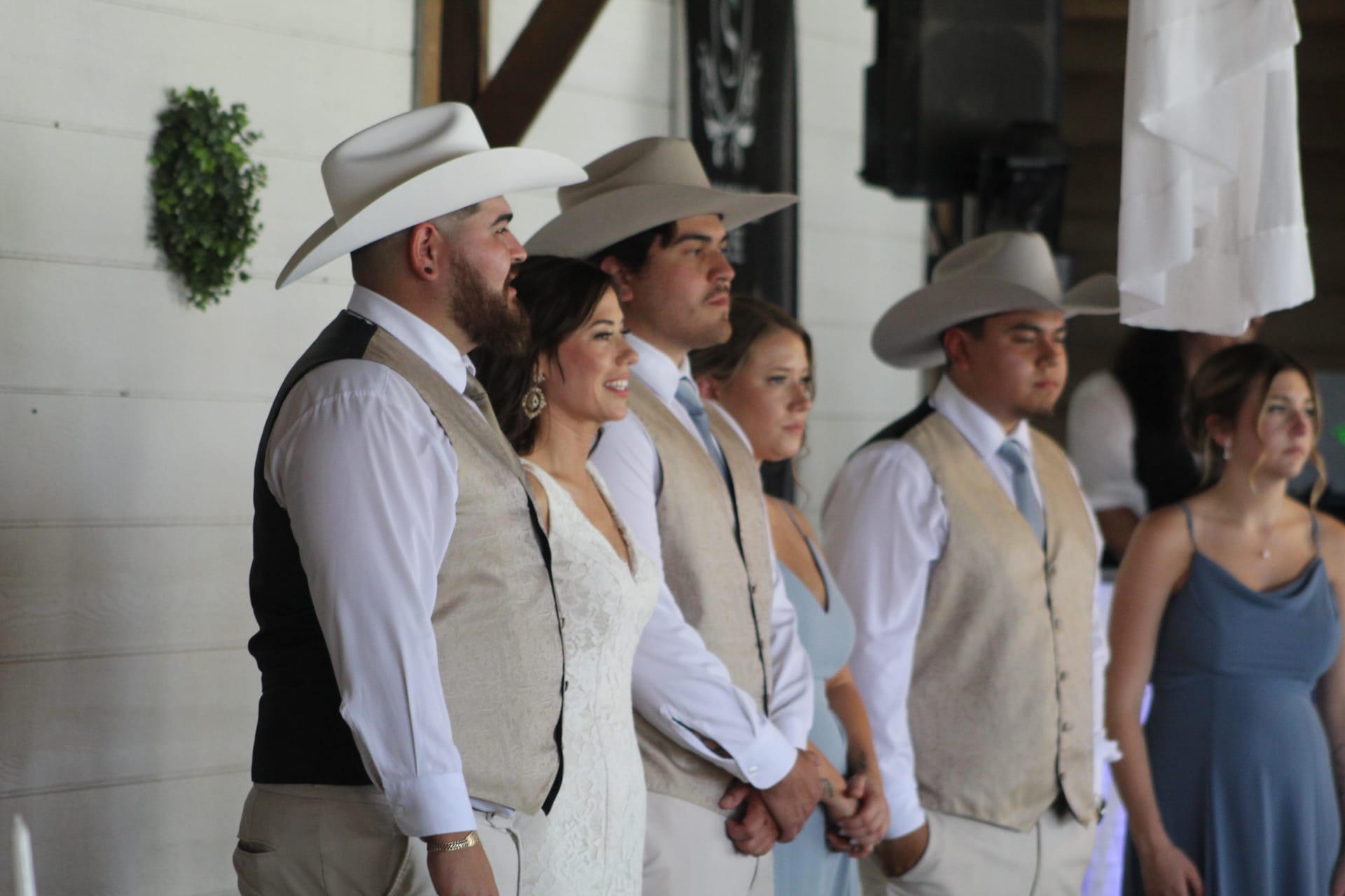 Wedding party in coordinated tan vests and white cowboy hats watching the ceremony inside a rustic white barn venue.