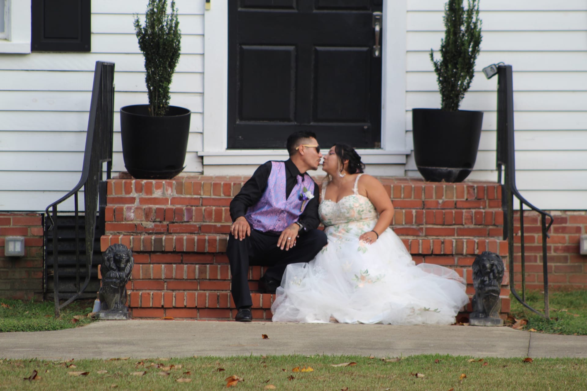 Bride in a floral ballgown and groom in a black suit with purple vest share a kiss while seated on the brick front steps of the historic Scarborough House estate.