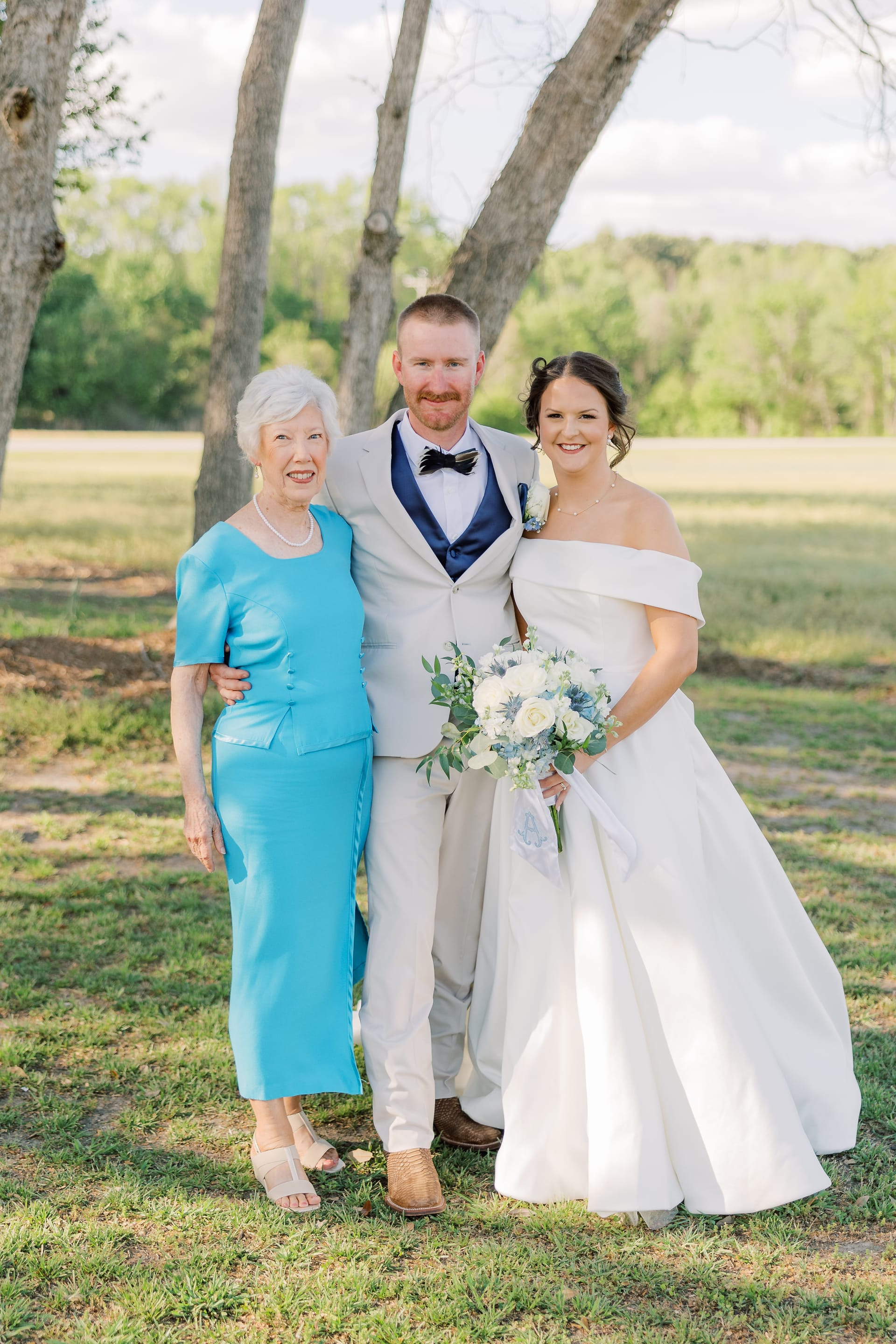 Bride in an off-shoulder white gown and groom in a light gray suit pose with an elderly woman in a turquoise dress beneath bare oak trees at Scarborough House.