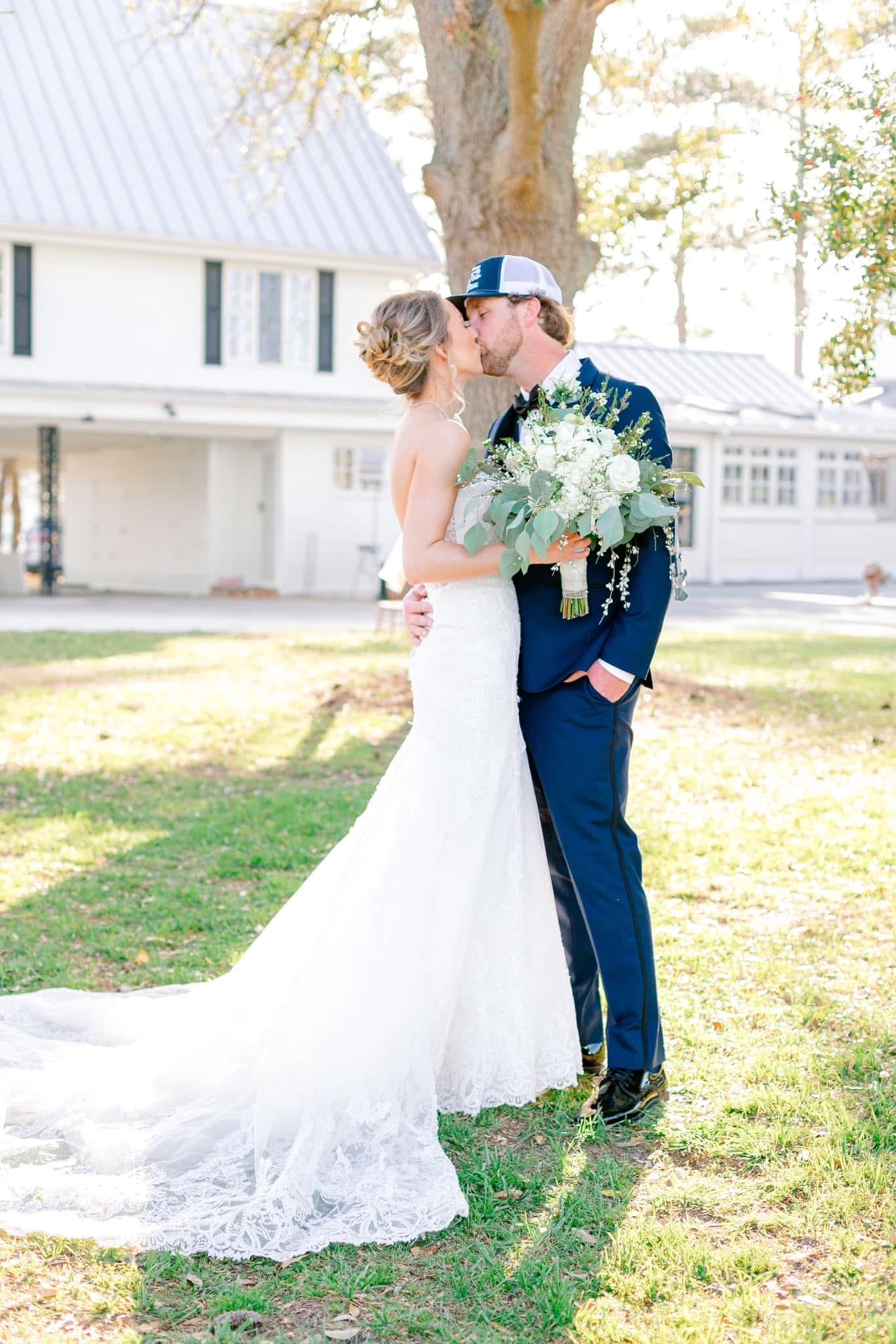 Bride in fitted lace gown and groom in navy suit sharing a kiss on the sunlit lawn in front of the historic 1821 Scarborough House manor.