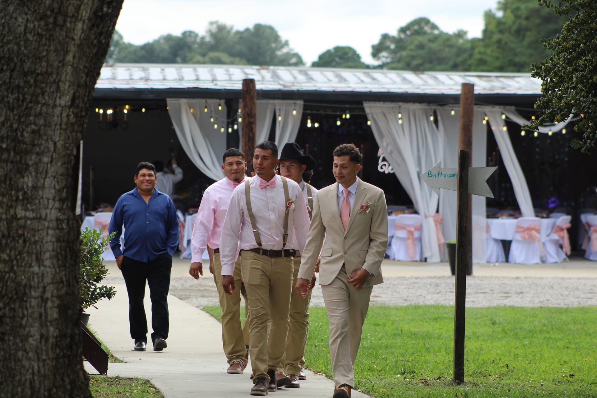 Groom in tan suit and groomsmen in pink shirts with suspenders walk along a pathway toward the camera with the decorated Scarborough House magnolia pavilion reception venue in the background.