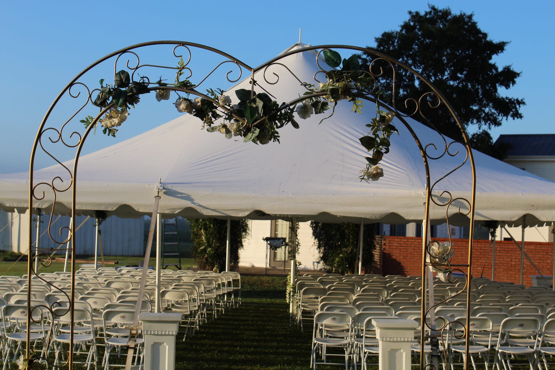 Ornate wrought-iron wedding arch adorned with white roses and greenery frames a sunlit outdoor ceremony aisle lined with white folding chairs at Scarborough House historic estate.
