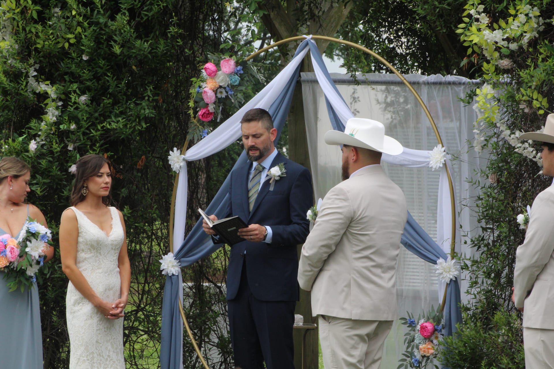 Bride in white lace gown and groom in tan suit with white cowboy hat exchange vows beneath a circular gold arch draped in dusty blue fabric and colorful florals on the ceremony lawn.