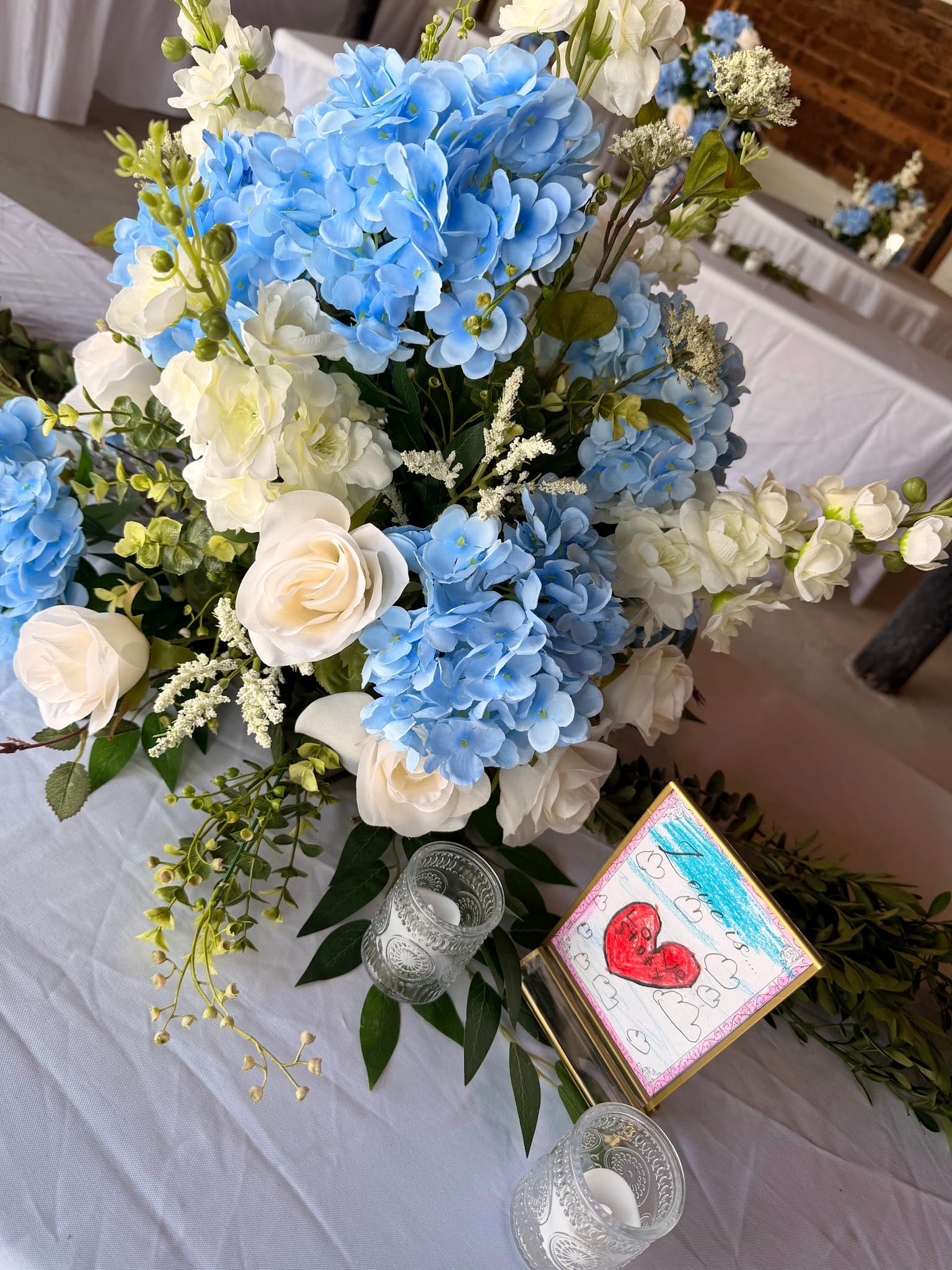 Lush wedding centerpiece of blue hydrangeas and white roses on a white linen table with crystal votive candles and a hand-drawn heart card at Scarborough House.