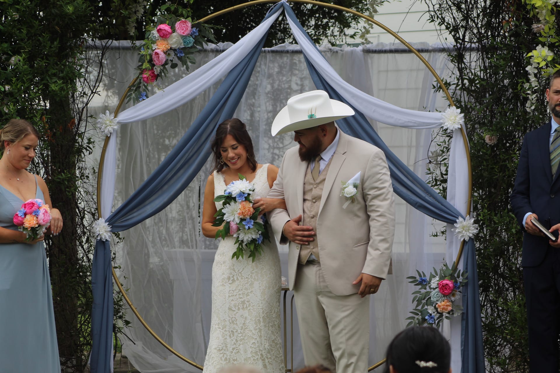 Bride in lace gown and groom in tan suit and white cowboy hat share a joyful moment beneath a gold circular arch draped in dusty blue fabric and colorful florals during an outdoor wedding ceremony.