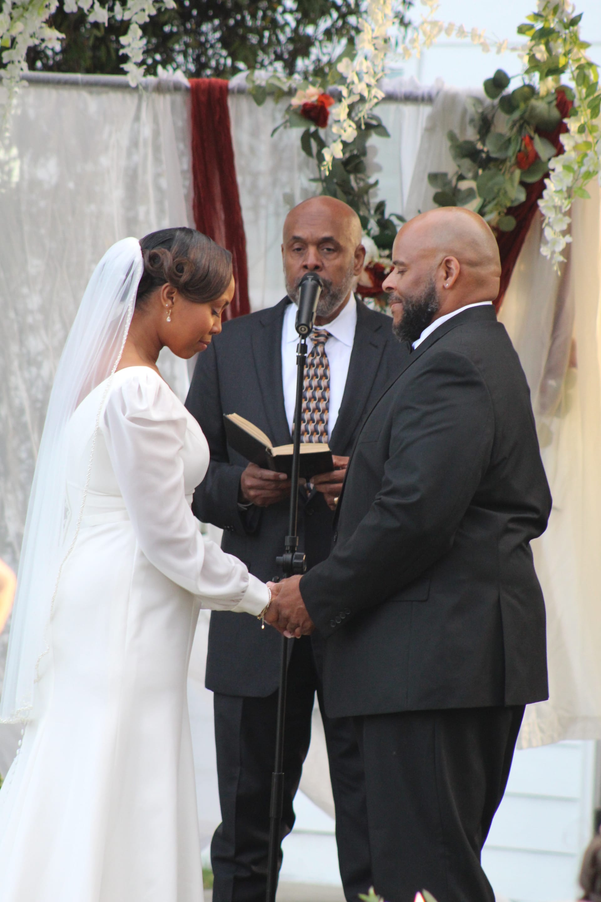 Bride in a long-sleeve white gown and groom in a black tuxedo hold hands during their outdoor wedding vow exchange as the officiant reads from a book beneath a floral and greenery arch.