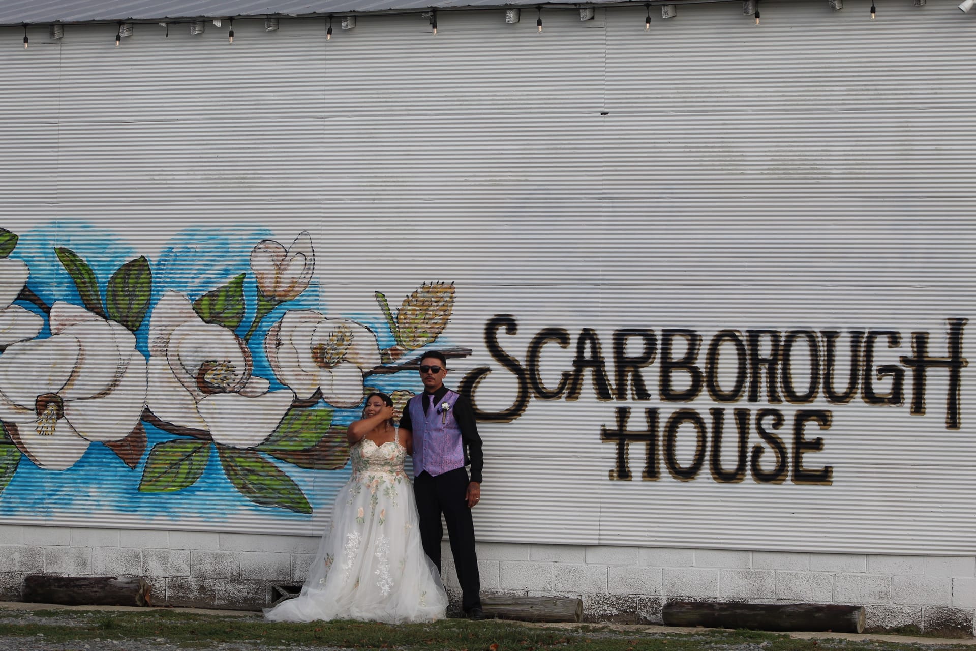 Bride in a floral ballgown and groom in a dark suit with purple vest pose together in front of the Scarborough House magnolia mural painted on a corrugated metal exterior wall.