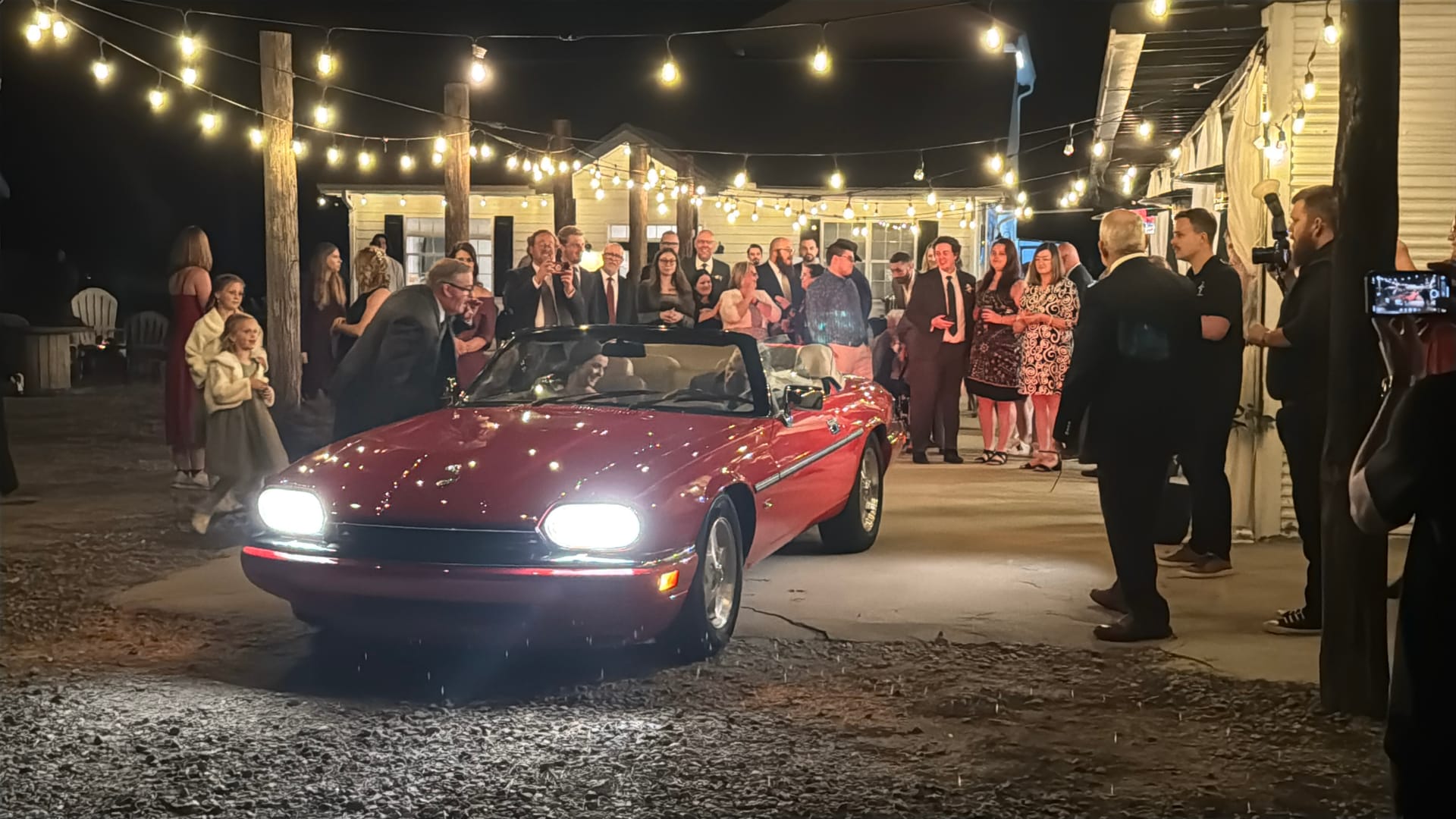 Guests gathered under festoon lights cheering as a couple departs in a red classic convertible at a nighttime wedding reception at Scarborough House.