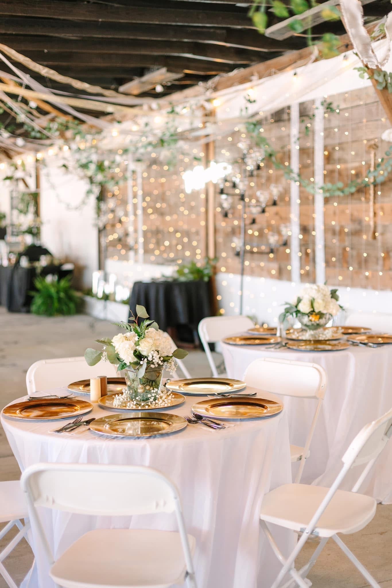 Elegantly set round reception tables with white linens, gold charger plates, and white floral centerpieces surrounded by warm fairy lights and greenery at Scarborough House historic estate wedding venue.