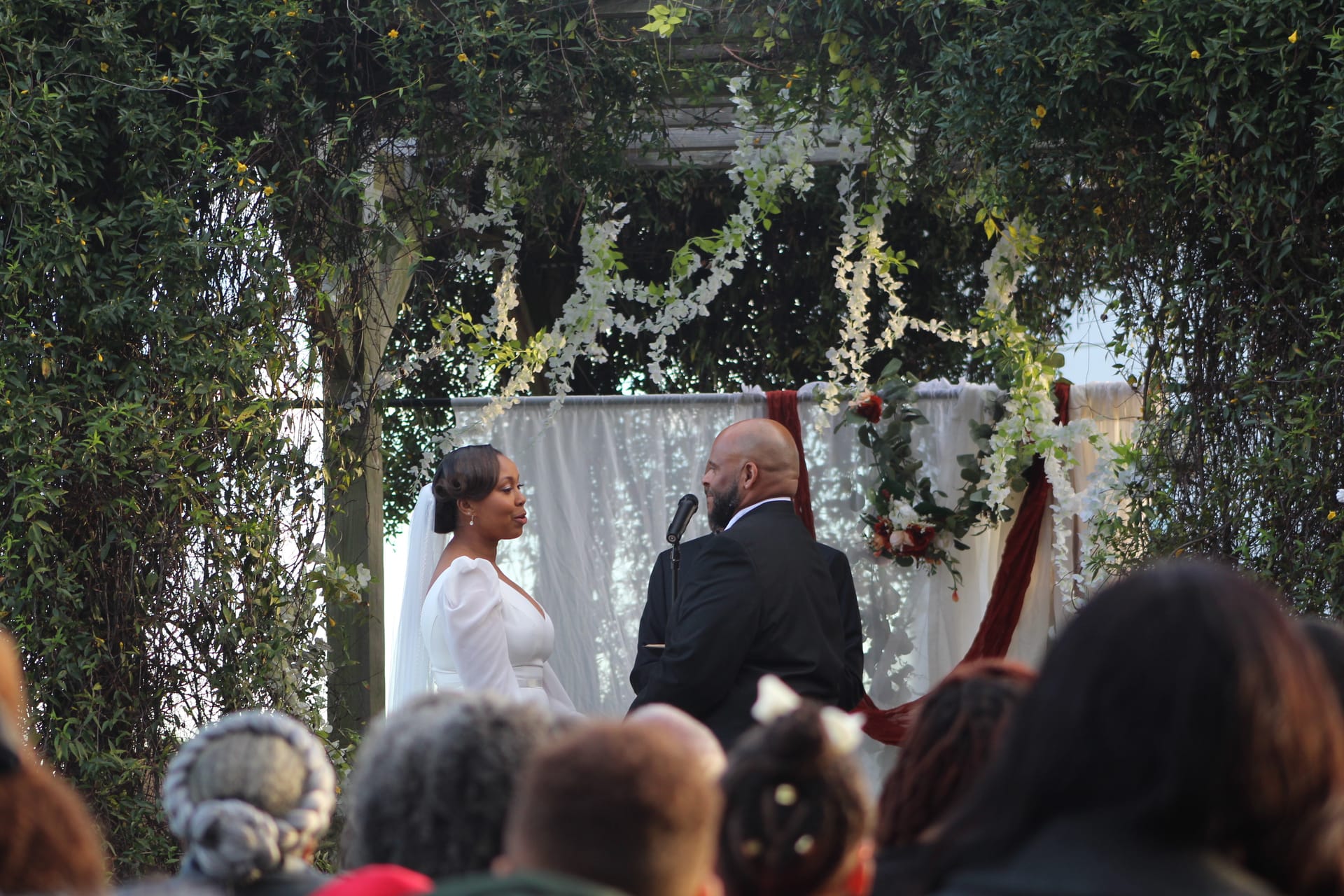 Bride in a white gown and groom in a black suit exchange vows beneath a floral-draped pergola surrounded by lush greenery and white drapery at an outdoor garden wedding ceremony.