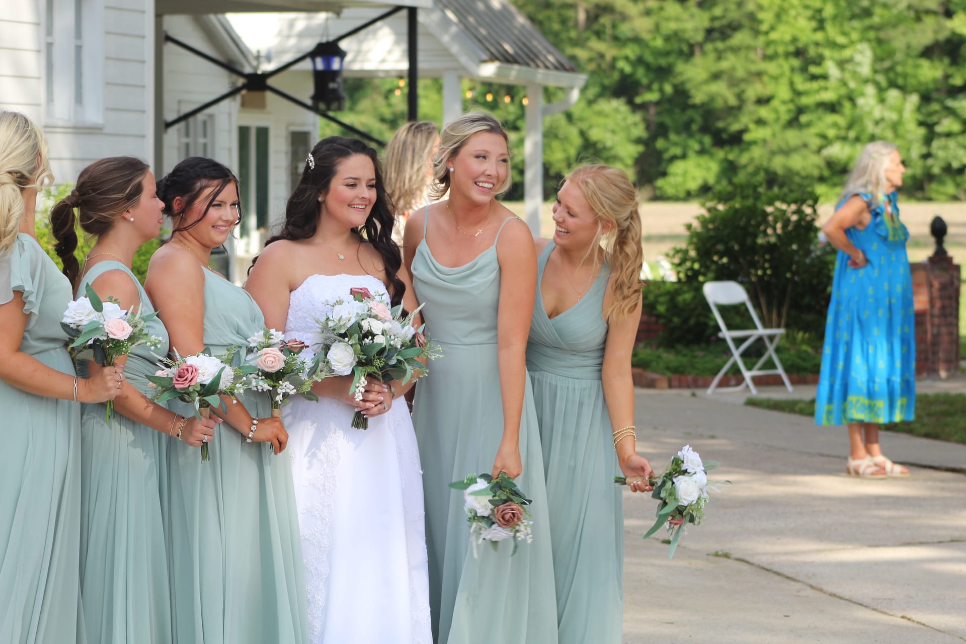 Bride in white lace gown laughs with her bridesmaids in sage green dresses holding pink and white floral bouquets outside the historic Scarborough House manor.