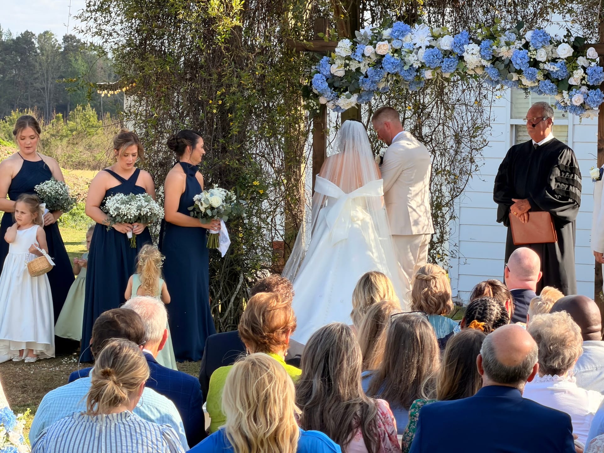 Bride and groom standing at a floral arch adorned with blue and white hydrangeas during an outdoor wedding ceremony at Scarborough House historic estate, surrounded by navy-clad bridesmaids and seated guests.